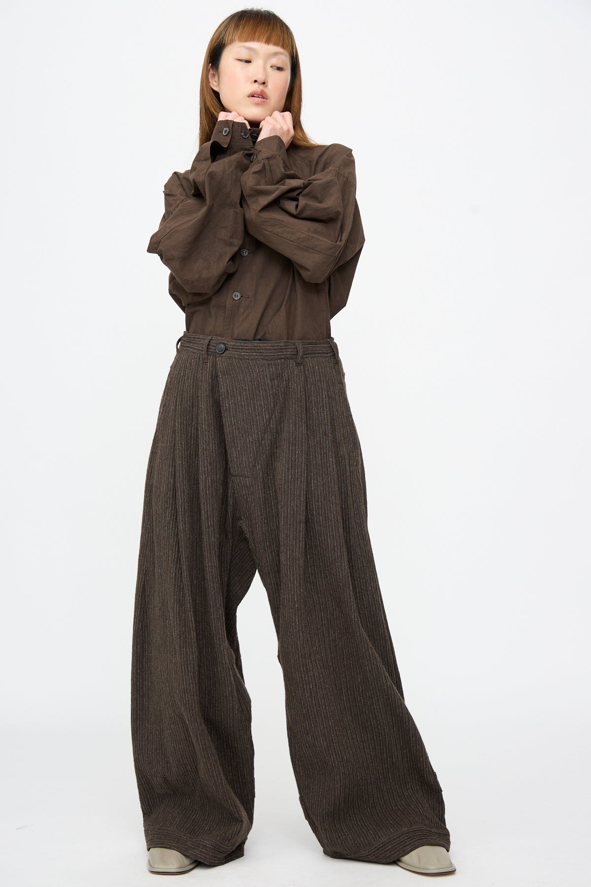 A person wearing a brown button-up shirt and Jan-Jan Van Essche Yak Linen Cotton Trousers #93 in Brown Striped, featuring a double pleated, extra wide fit with thin vertical stripes.