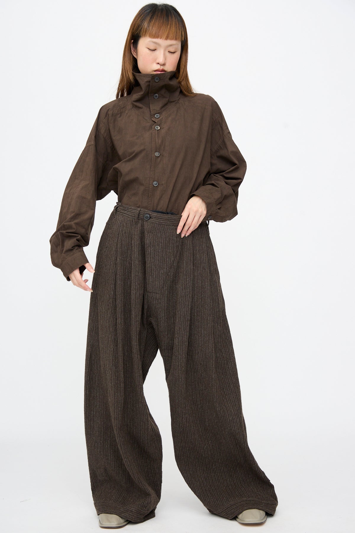 Person wearing a brown high-collar button-down shirt and Jan-Jan Van Essche Yak Linen Cotton Trousers #93 in Brown Striped, which are brown pinstriped double-pleated wide-leg trousers.