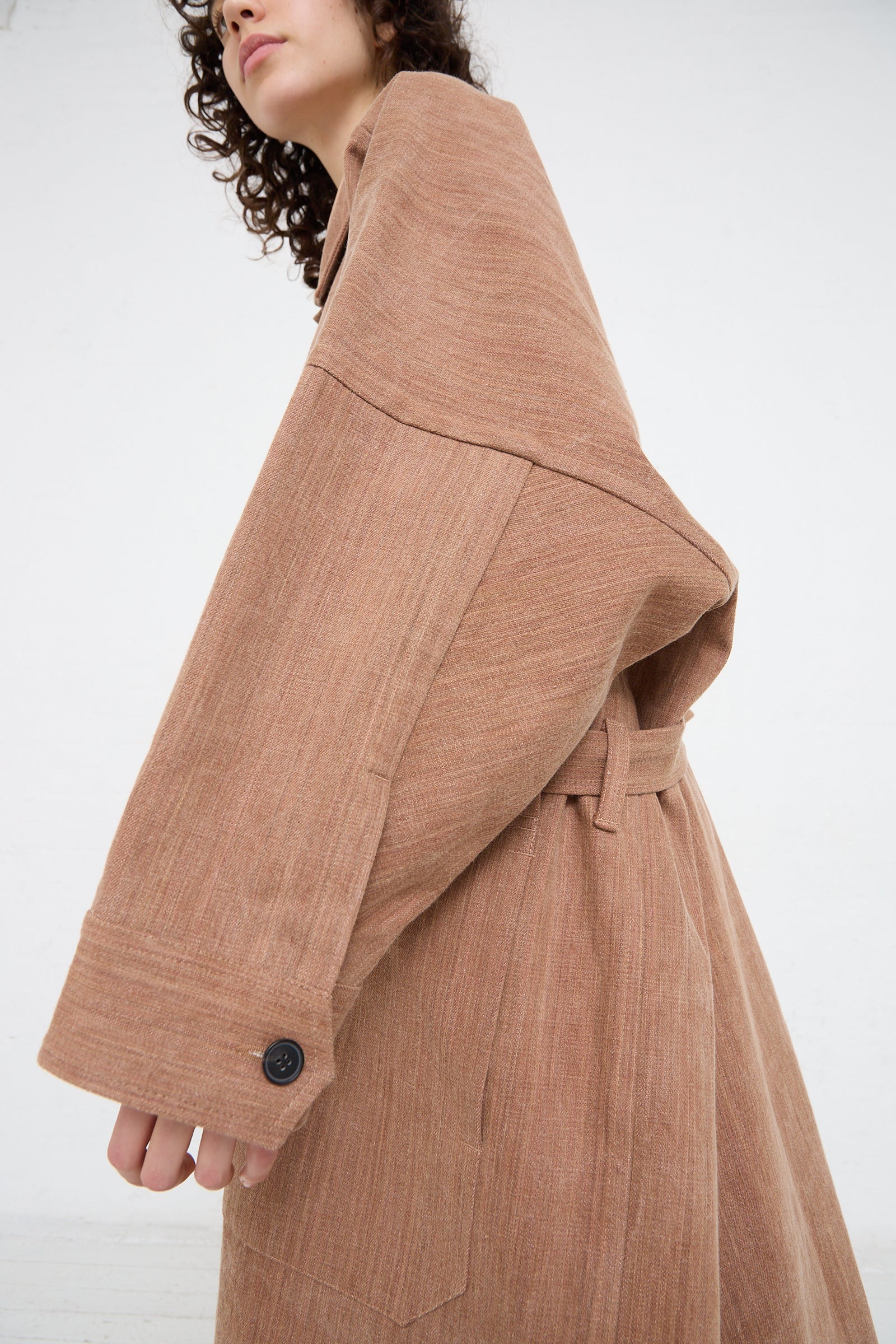 A woman in a brown trench coat with a self tie belt made of heavy Woven Cotton Long Coat in Kakishibu by Jan-Jan Van Essche. Side view.