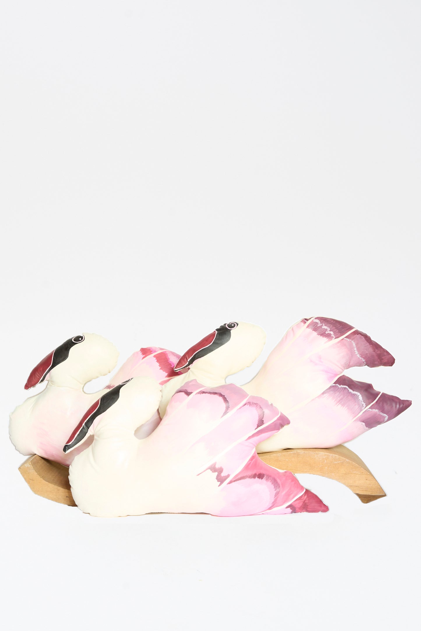 The Hand Painted Swan Pillow II by Jess Feury features three swans with pink and white feathers on a wooden base, arranged closely together to evoke handmade charm against a plain white background.