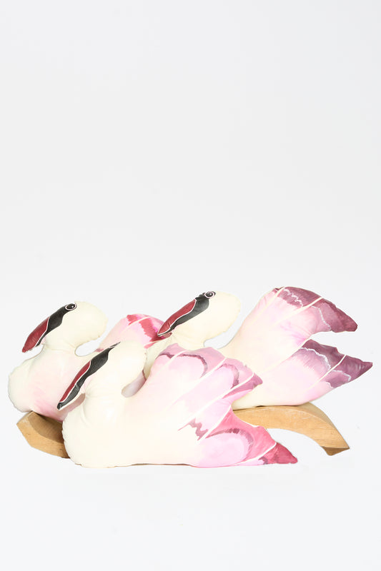 The Hand Painted Swan Pillow II by Jess Feury features three swans with pink and white feathers on a wooden base, arranged closely together to evoke handmade charm against a plain white background.