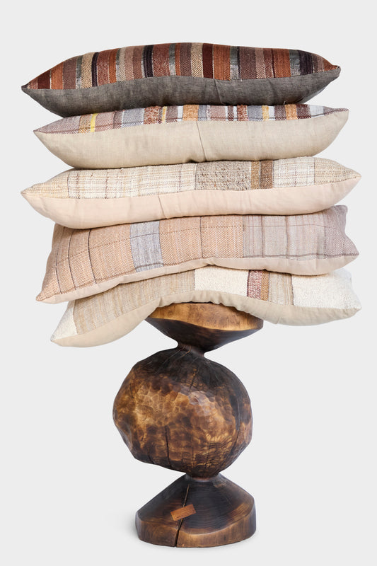 Six striped and textured throw pillows, including the Jess Feury Handmade Rock Slab Pillow in Neutral and Olive, are stacked on an abstract sculptural wooden base against a plain white background.