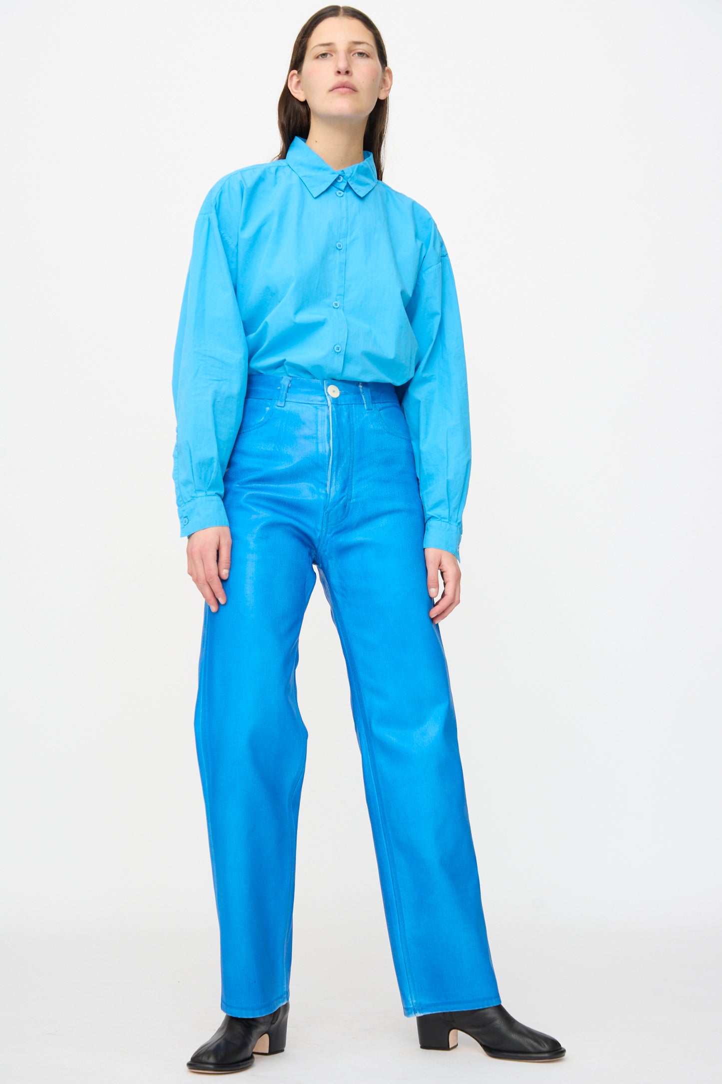 A person stands against a plain background wearing a light blue button-up shirt, Jesse Kamm 225 Jeans in Electric Blue Lacquer, and black heeled shoes.