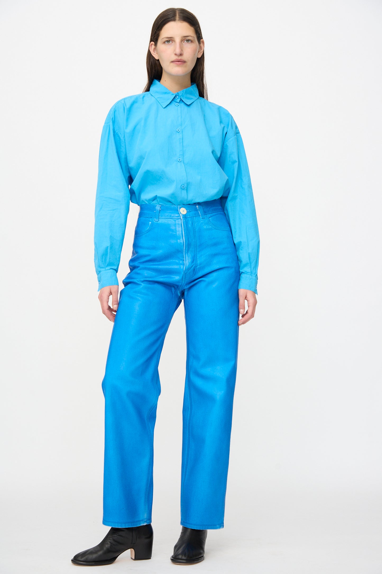 A person stands facing forward against a plain white background, wearing a light blue button-up shirt and Jesse Kamm 225 Jeans in Electric Blue Lacquer, paired with black heeled shoes.