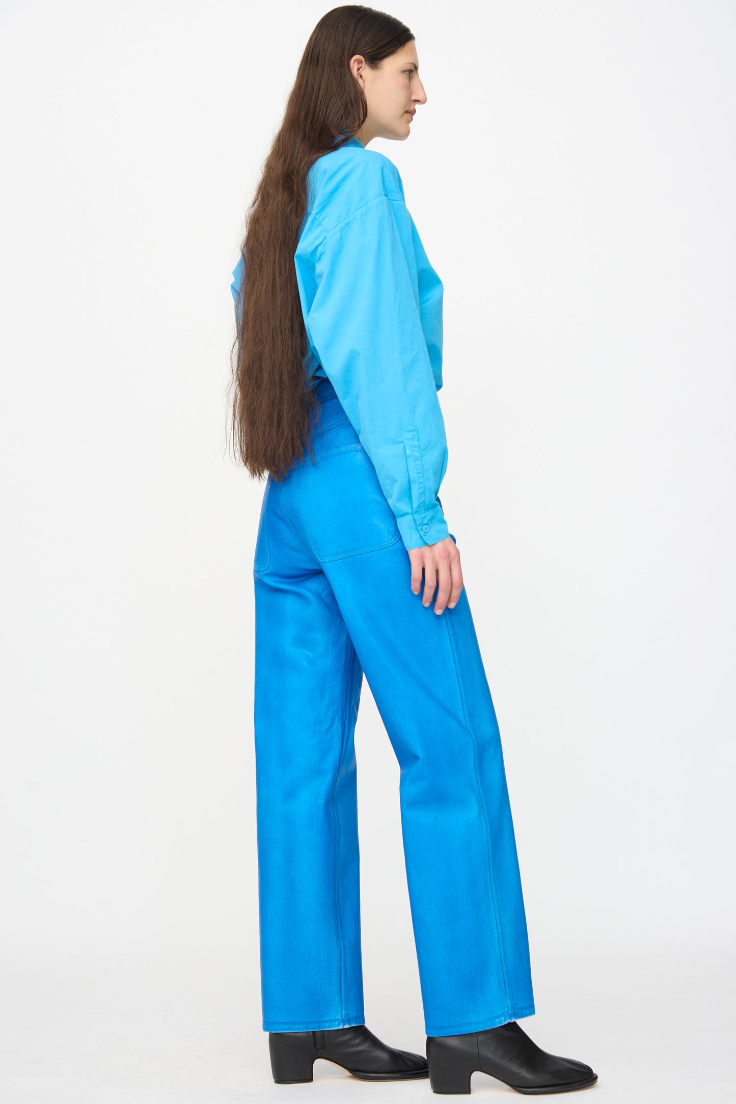 A person with long brown hair wears a light blue shirt, Jesse Kamm 225 Jeans in Electric Blue Lacquer, and black boots, standing sideways against a plain white background.