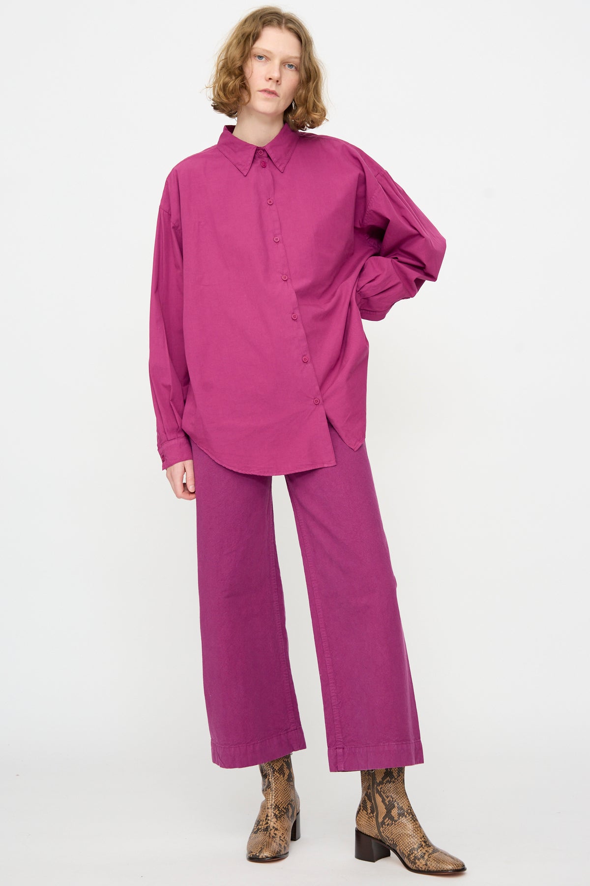 Person stands against a plain background wearing the Jesse Kamm Alberta Shirt in Beet, an oversized-fit artist shirt made from organic cotton, paired with matching wide-leg pants and brown snakeskin-patterned boots.