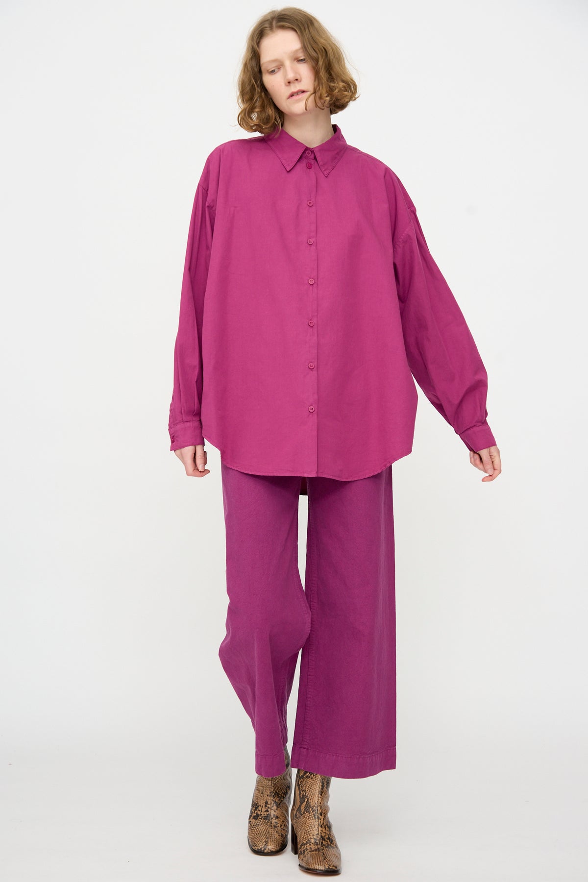 Wearing the Jesse Kamm Alberta Shirt in Beet—an oversized fit artist shirt crafted from organic cotton—paired with matching wide-leg pants and brown snakeskin boots, the person stands against a plain white background.