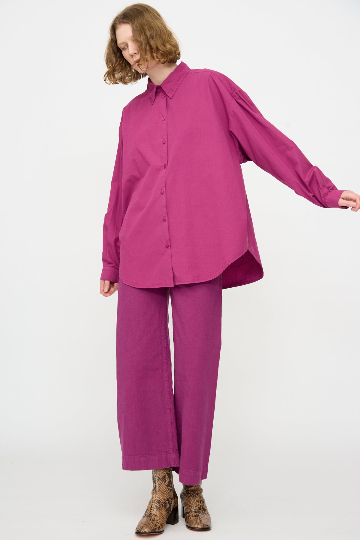 A person wearing the Jesse Kamm Alberta Shirt in Beet, styled as an oversized fit, with matching pants and tan snake-print boots, stands against a plain white background.
