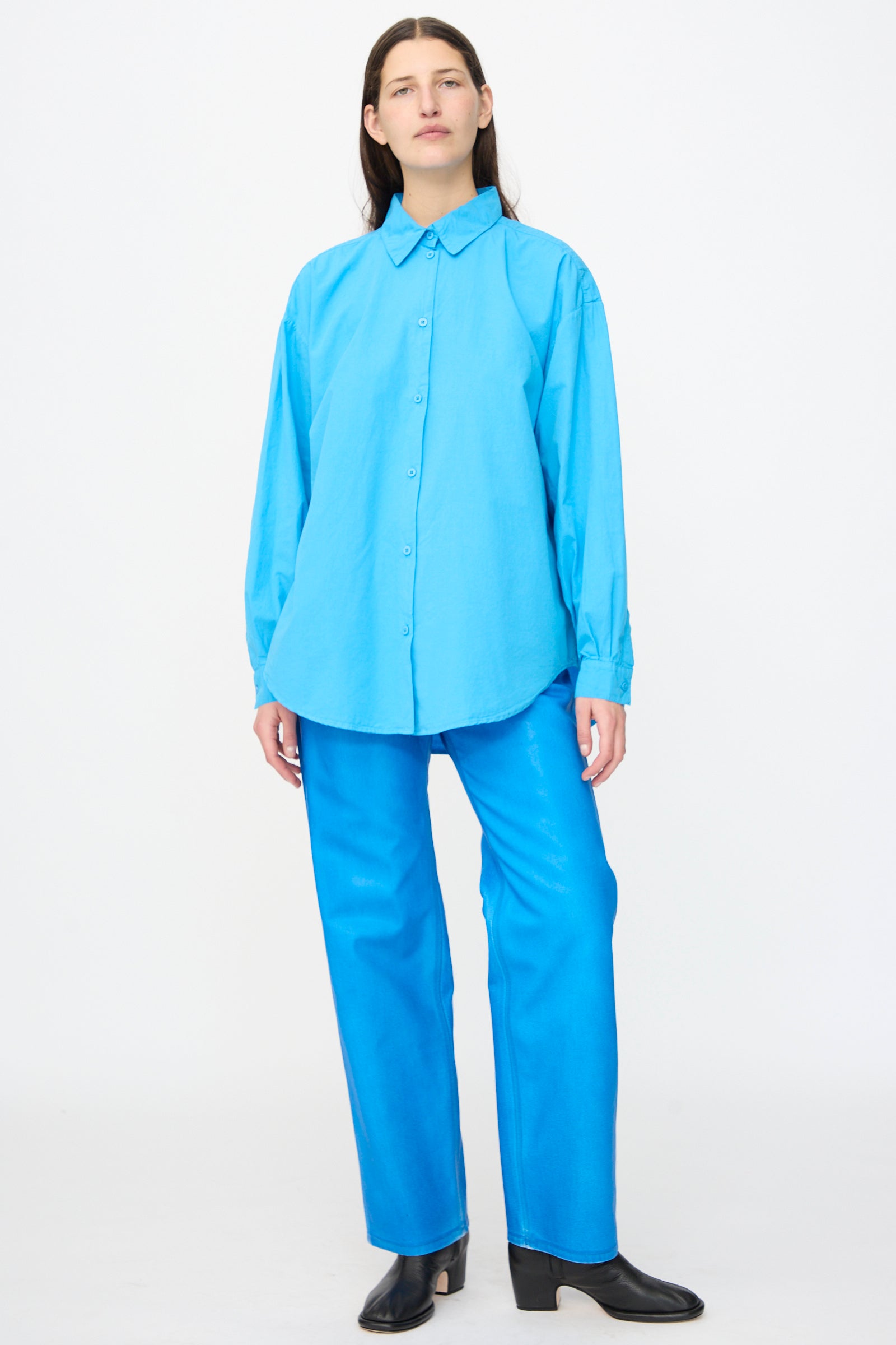 A person stands facing forward wearing the Jesse Kamm Alberta Shirt in Pool Blue, paired with matching blue pants and black heeled shoes against a plain white background.