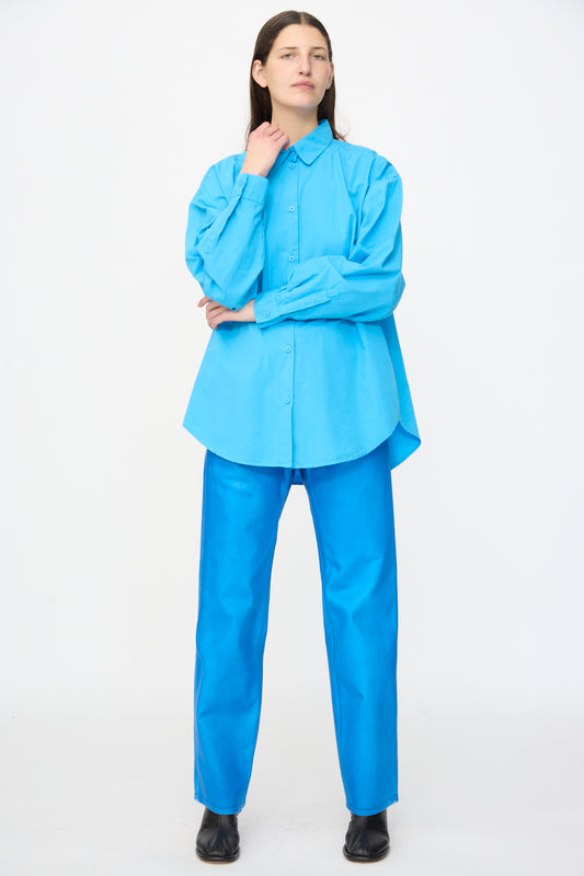 A person stands against a white background wearing the Jesse Kamm Alberta Shirt in Pool Blue, paired with bright blue pants and black shoes.