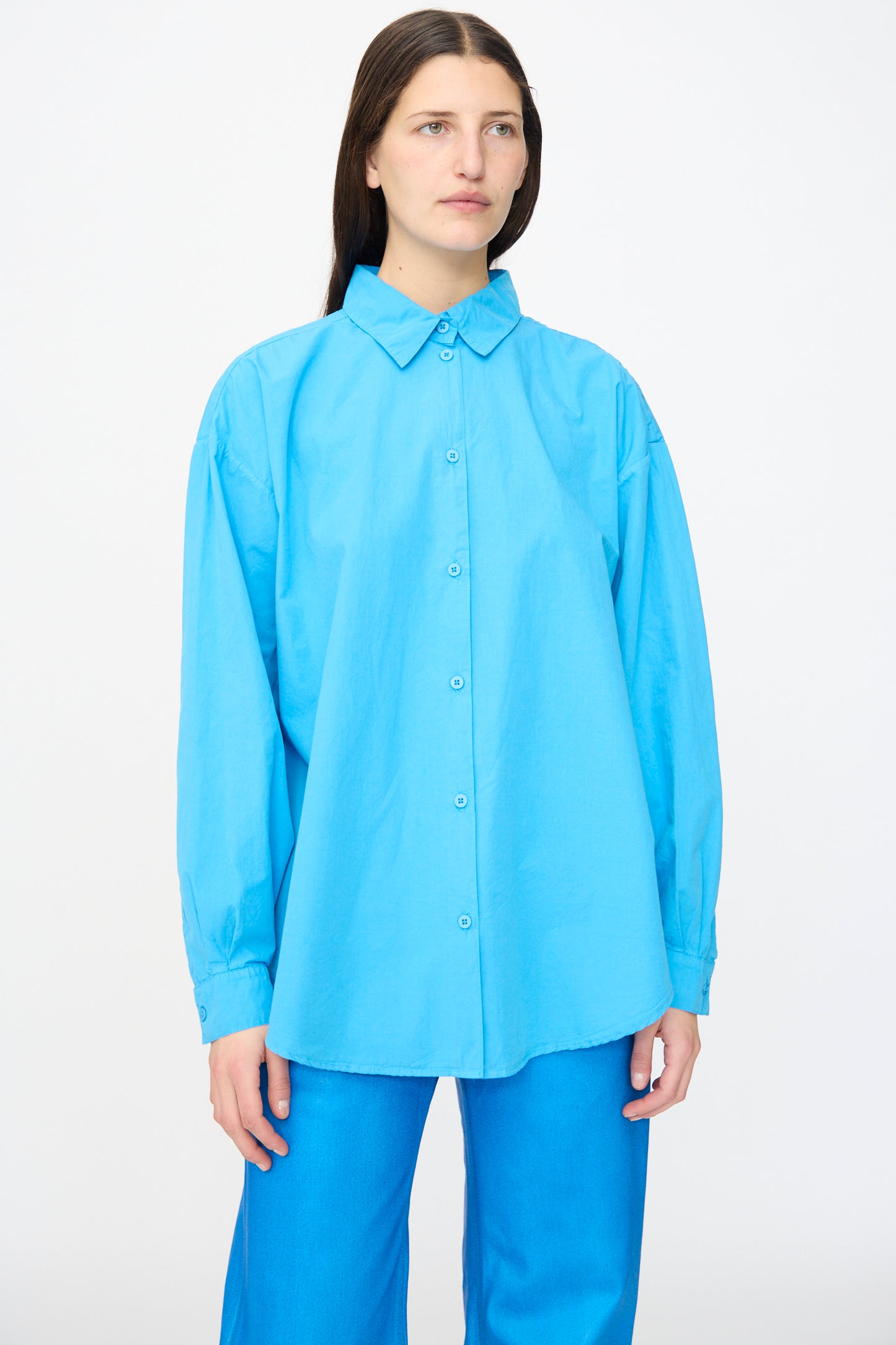 A woman stands against a plain white background wearing the Jesse Kamm Alberta Shirt in Pool Blue, a loose-fitting, long-sleeved organic cotton button-up, paired with blue pants.