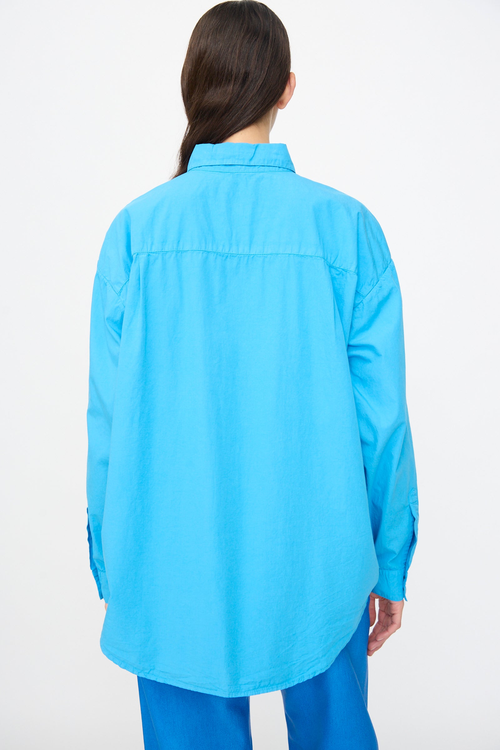 Model with long dark hair, seen from the back against a white background, wears the Jesse Kamm Alberta Shirt in Pool Blue—an oversized organic cotton design—paired with blue pants.