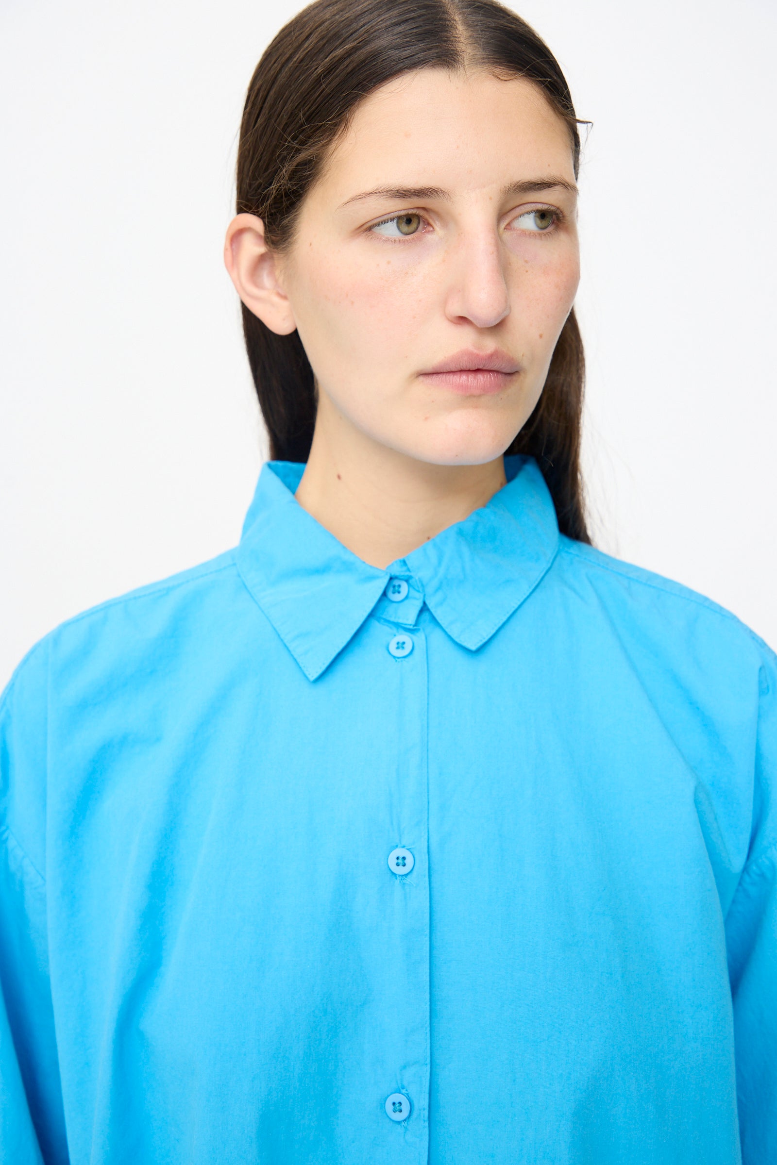 A person with straight brown hair wears the Jesse Kamm Alberta Shirt in Pool Blue, a loose-fitting organic cotton button-up, while looking slightly to the side against a plain white background.