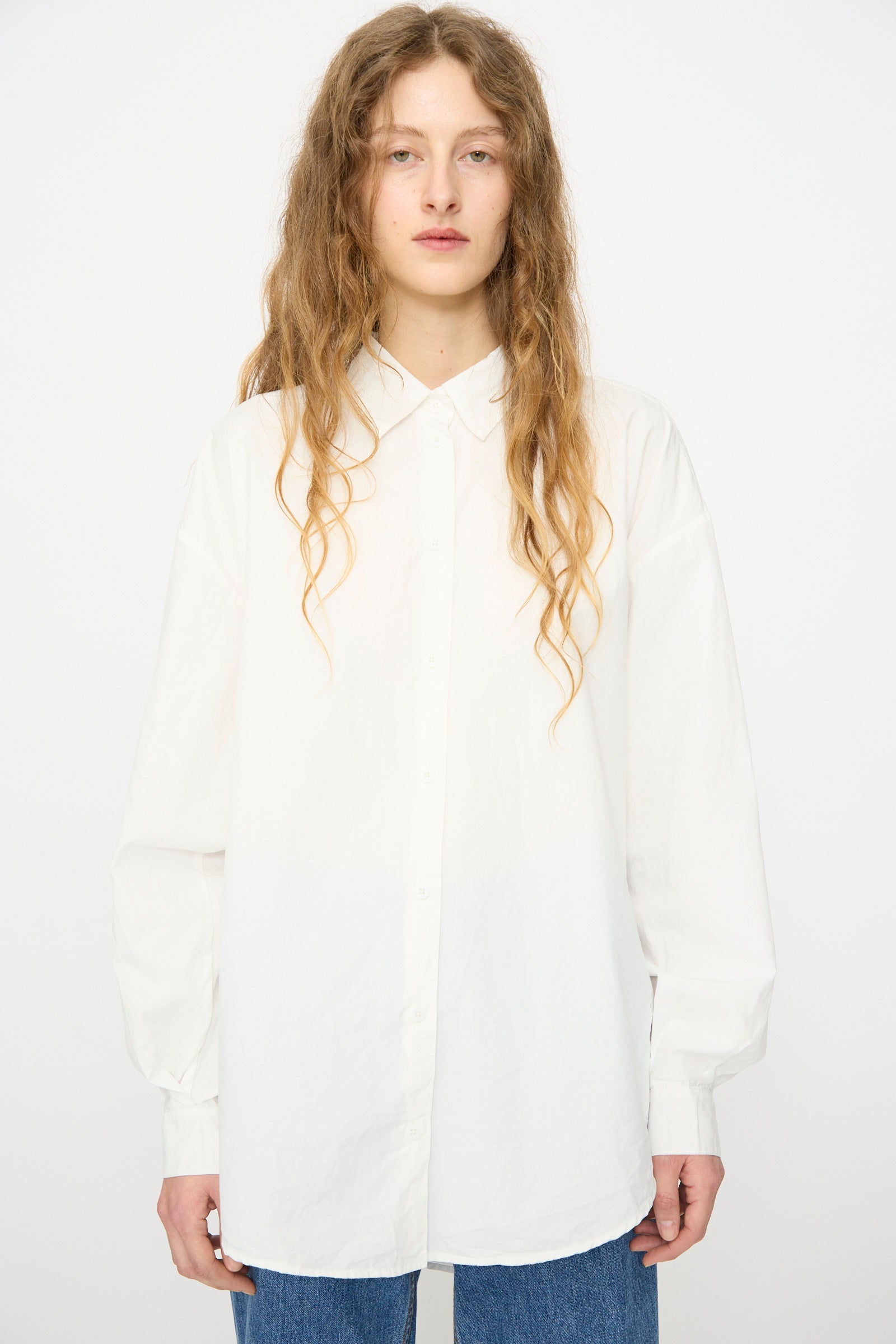 A person with long wavy hair wears the Jesse Kamm Alberta Shirt in Salt White, styled oversized with blue jeans, standing against a plain white background.