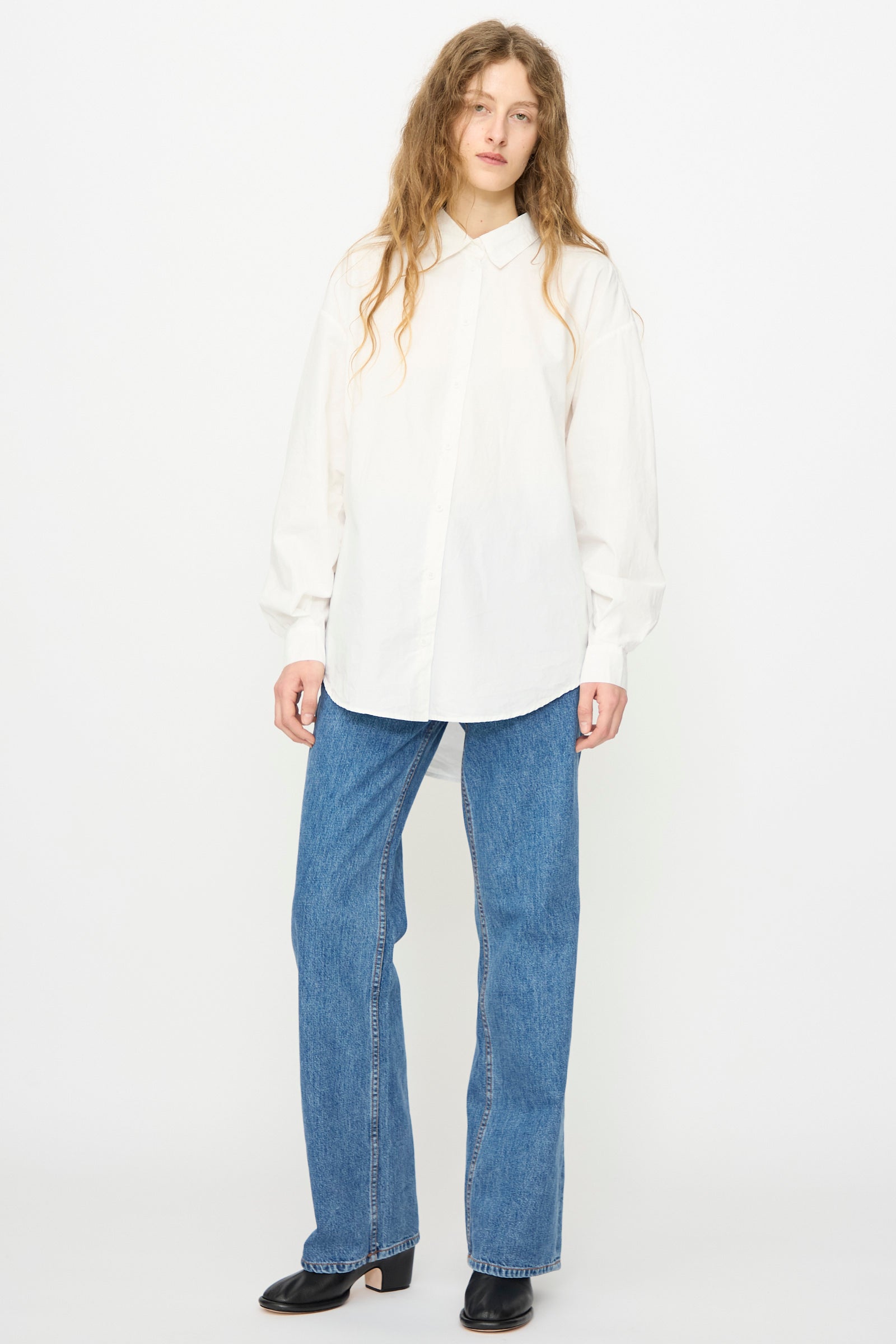 A person with long wavy hair wears the Jesse Kamm Alberta Shirt in Salt White, straight-leg blue jeans, and black shoes, standing against a plain white background.