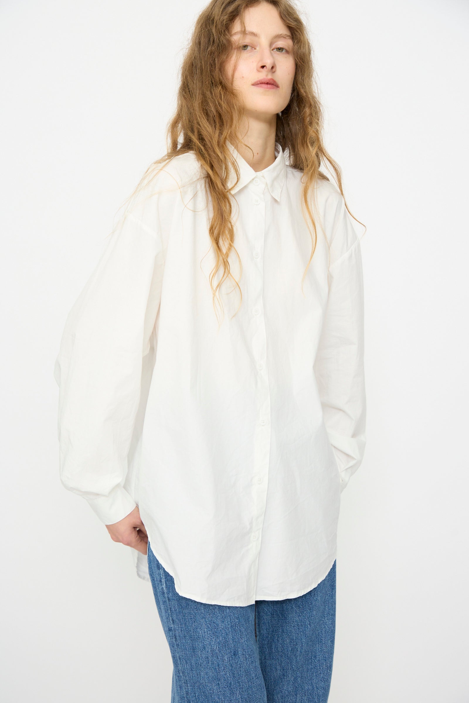 A person with long wavy hair wears the Jesse Kamm Alberta Shirt in Salt White and blue jeans, standing against a plain white background.