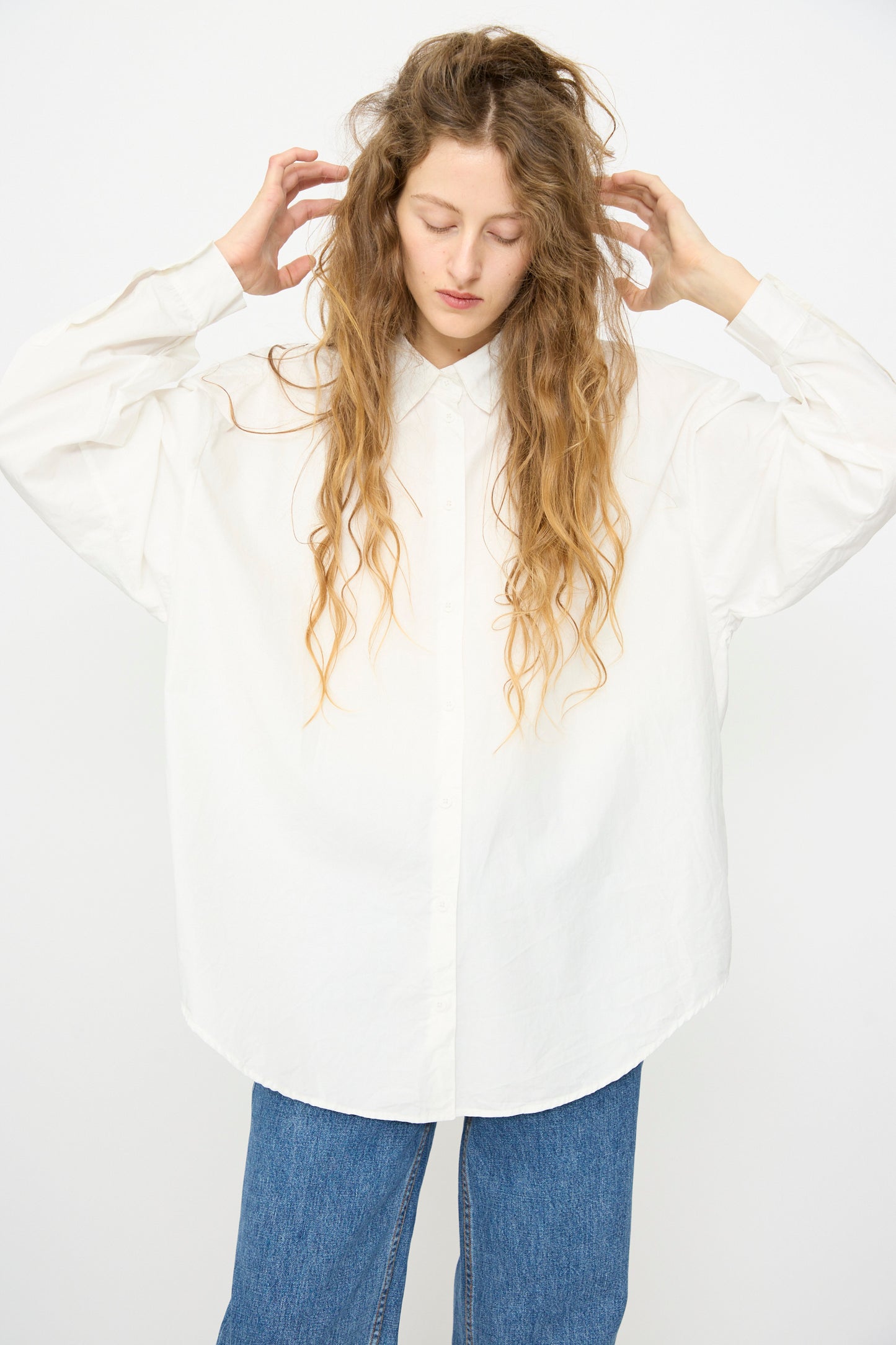 A person with long wavy hair wears the Jesse Kamm Alberta Shirt in Salt White and blue jeans, standing against a plain white background.