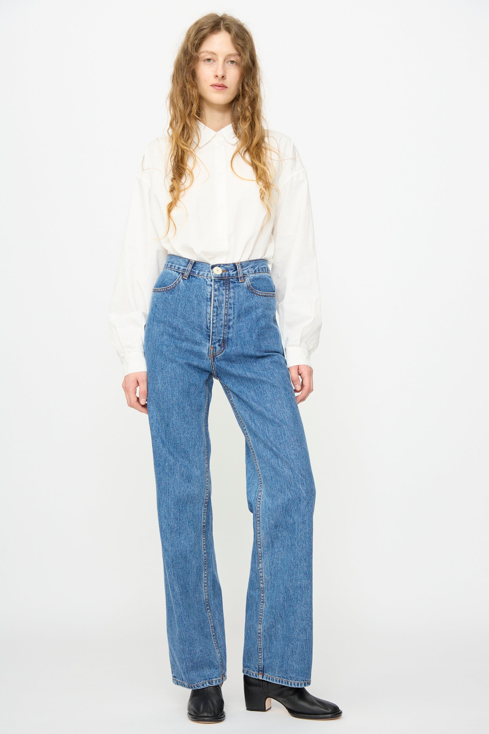 A person with long wavy hair wears a white button-up shirt, Jesse Kamm's Alta Jean in Cowboy Blue (high waisted, regenerative Japanese denim, boot cut), and black heeled ankle boots, standing against a plain white background.