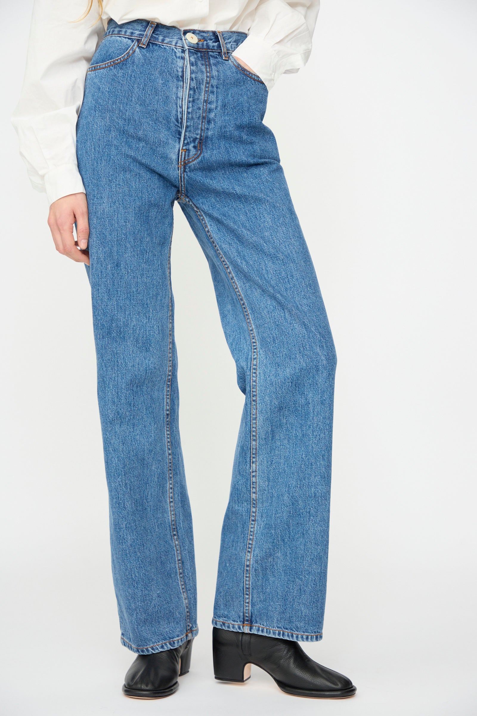 Wearing Jesse Kamm’s Alta Jean in Cowboy Blue—a high waisted, straight-leg jean in regenerative Japanese denim—with a white long-sleeve shirt and black heeled boots, one hand in pocket, posed against a plain background.