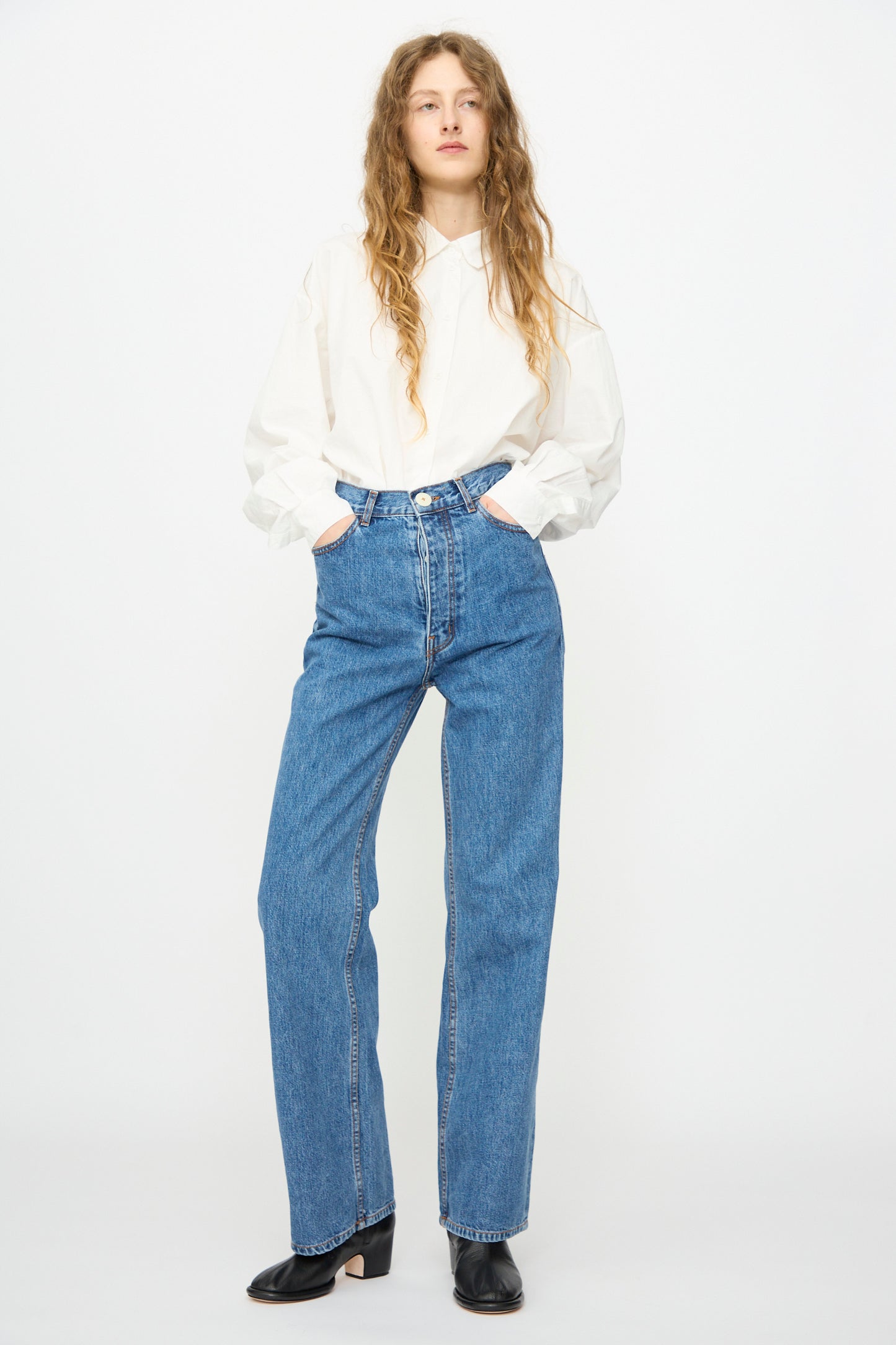 A person stands against a white background, wearing Jesse Kamm’s Alta Jean in Cowboy Blue—high-waisted jeans made from regenerative Japanese denim—paired with a loose white shirt, black shoes, and hands in pockets.