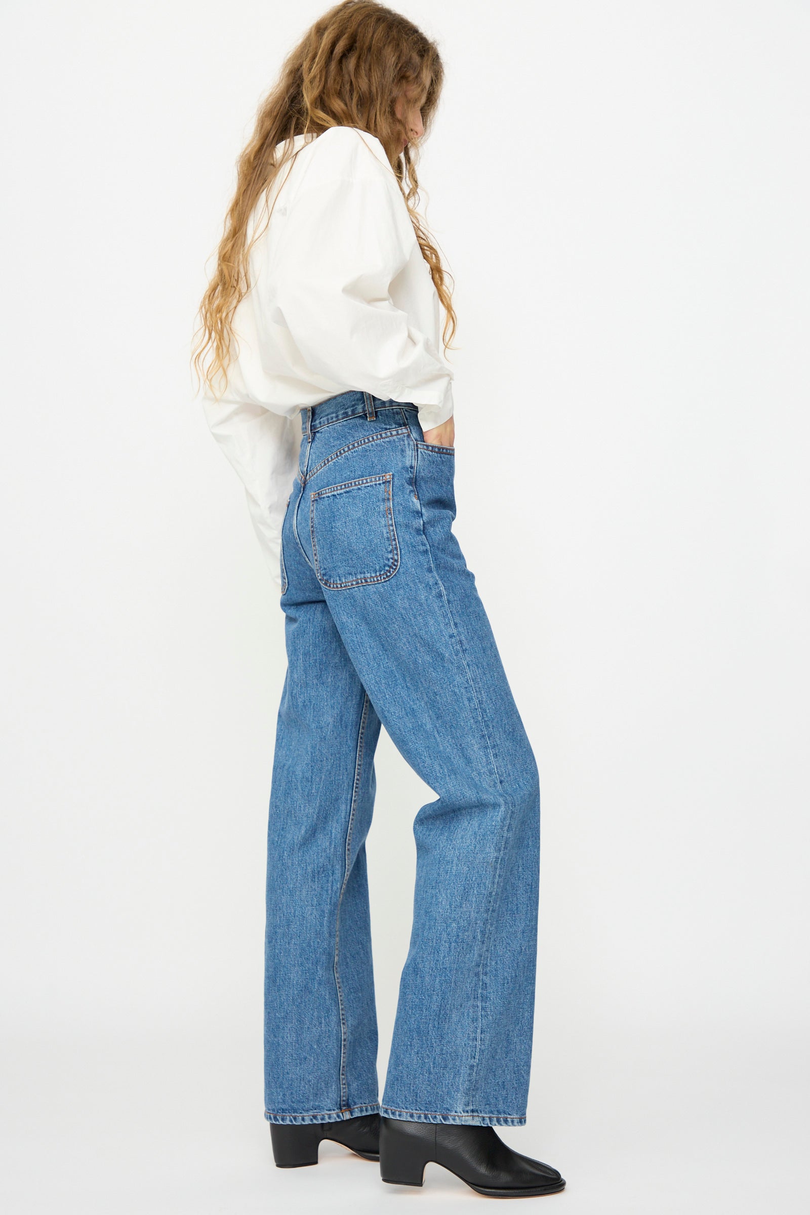 A person wearing a white long-sleeve shirt, Jesse Kamm Alta Jean in Cowboy Blue with a high waist and boot cut leg, and black boots, shown in side profile against a plain white background.