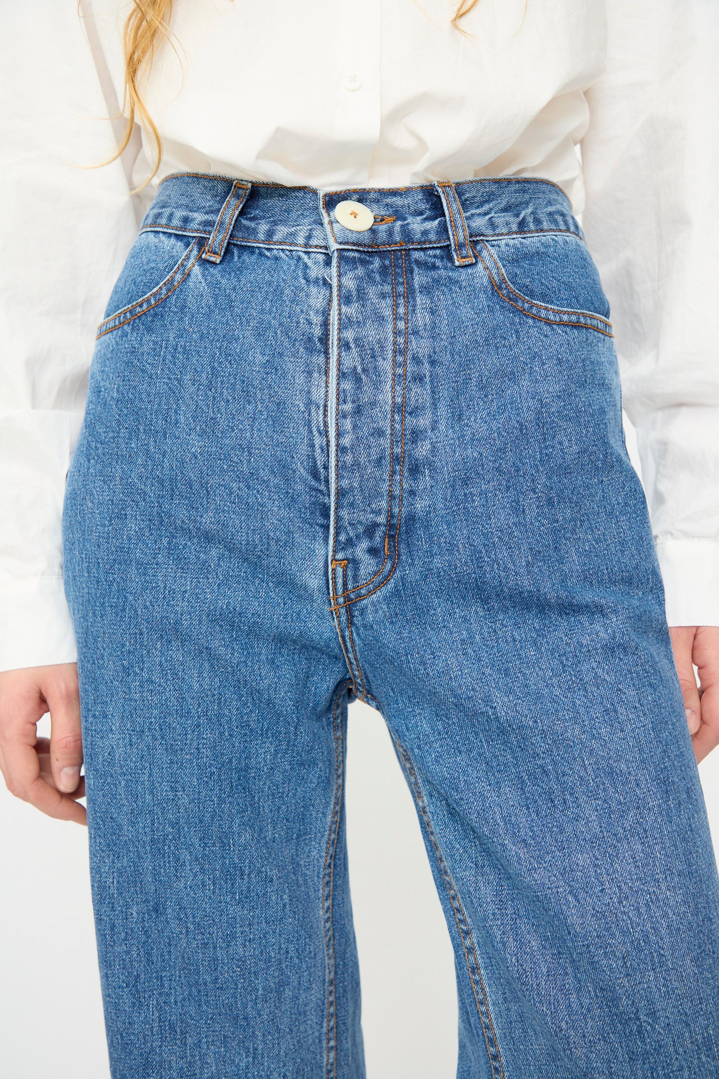 A person wears the Jesse Kamm Alta Jean in Cowboy Blue—high-waisted jeans made from regenerative Japanese denim—paired with a white button-up shirt, shown from chest to mid-thigh.