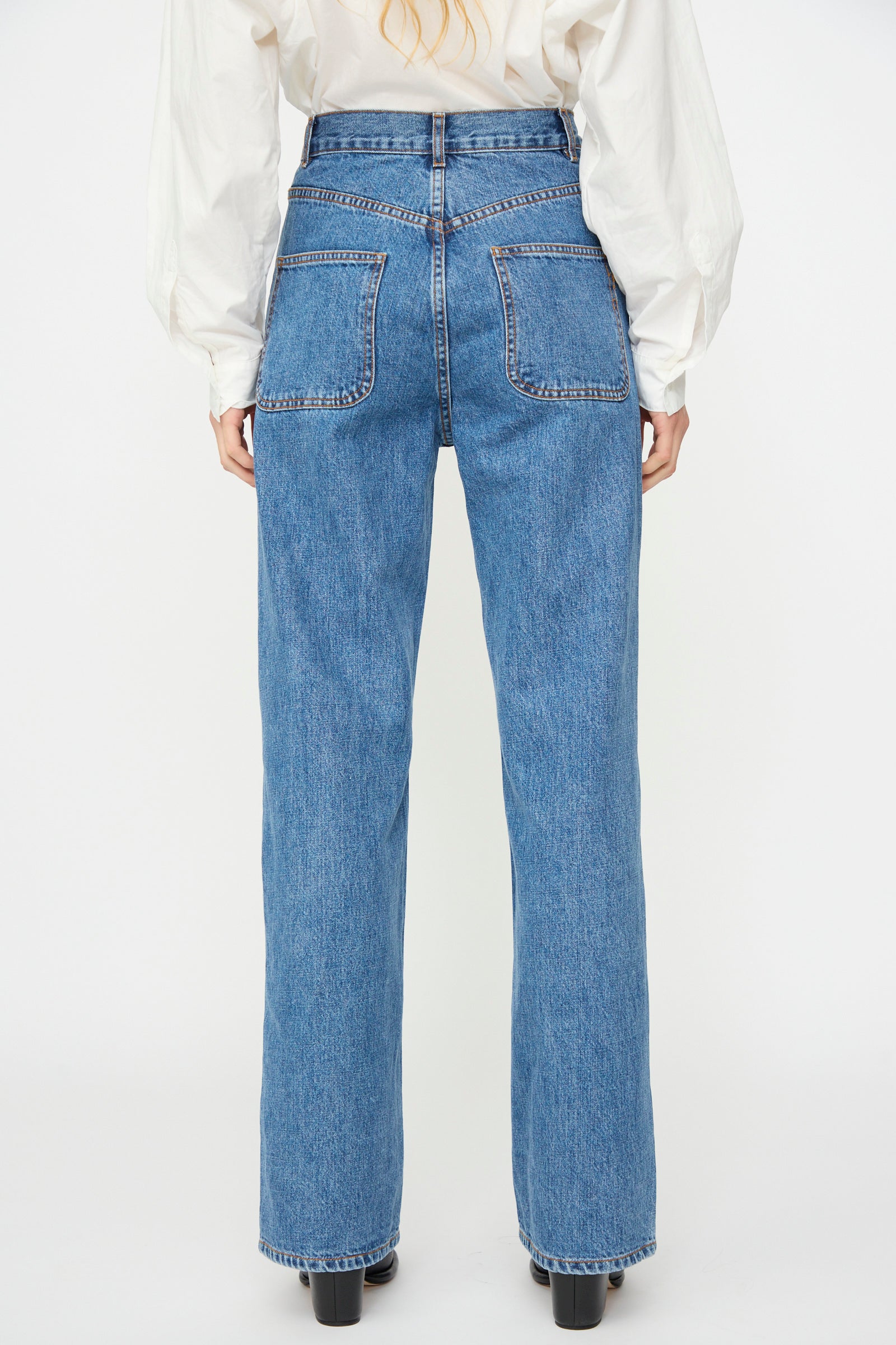 Model wears Jesse Kamm Alta Jean in Cowboy Blue—high-waisted, straight-leg jeans made from regenerative Japanese denim—and a white long-sleeve shirt, shown from the back against a white background.