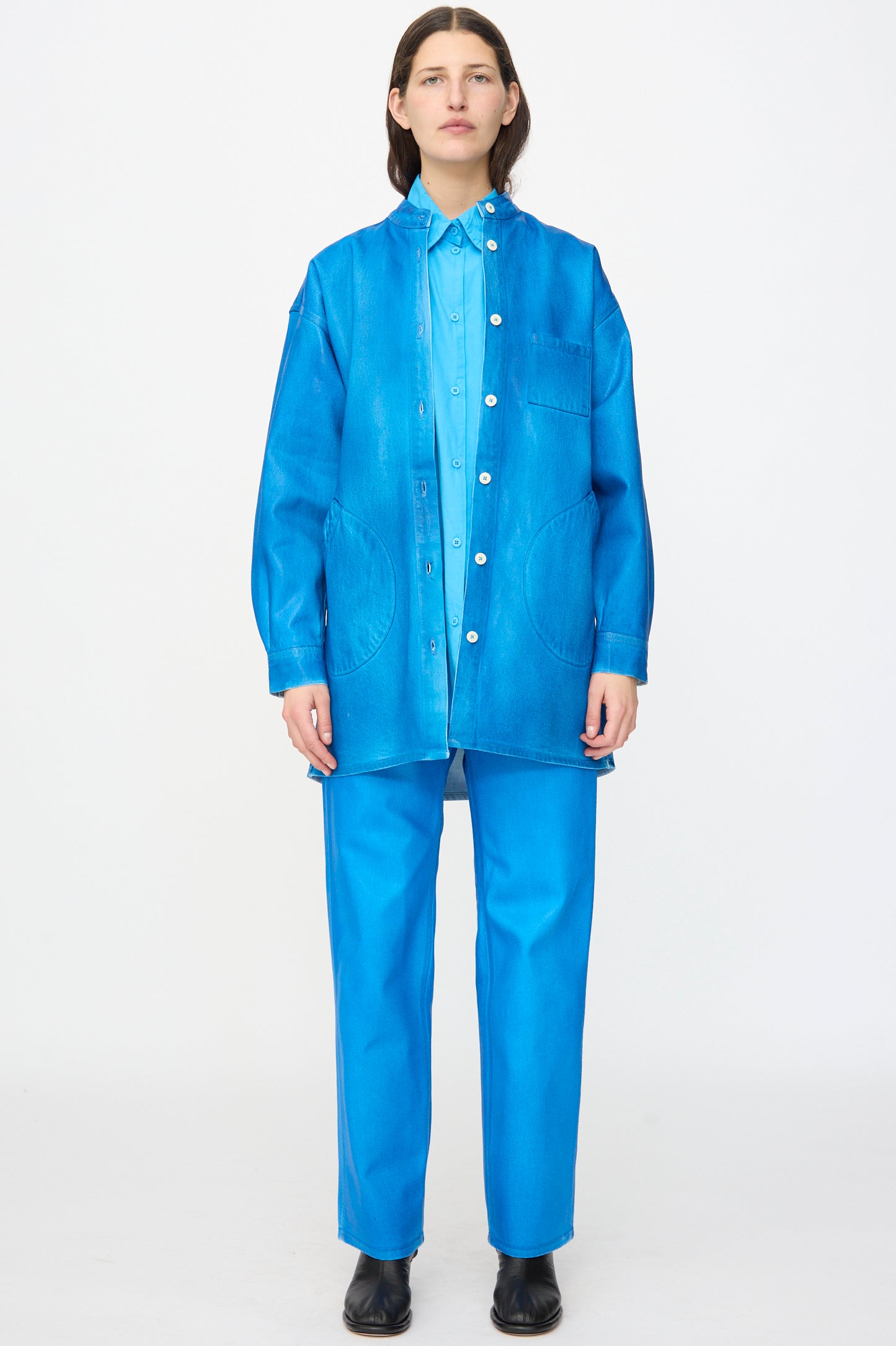 A person stands facing forward, wearing the Jesse Kamm Band Collar Okuda Jacket in Electric Blue Lacquer with loose pants and black shoes against a plain white background.