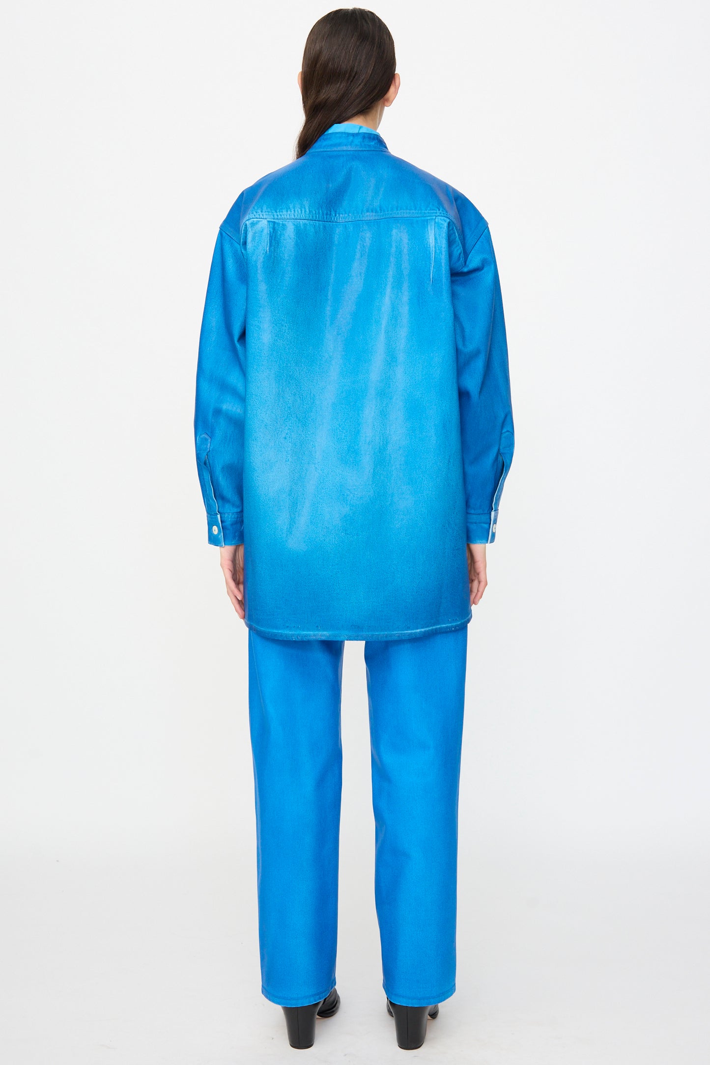 A person with their back to the camera wears the Jesse Kamm Band Collar Okuda Jacket in Electric Blue Lacquer, crafted from regenerative Japanese denim, paired with matching pants, against a plain white background.
