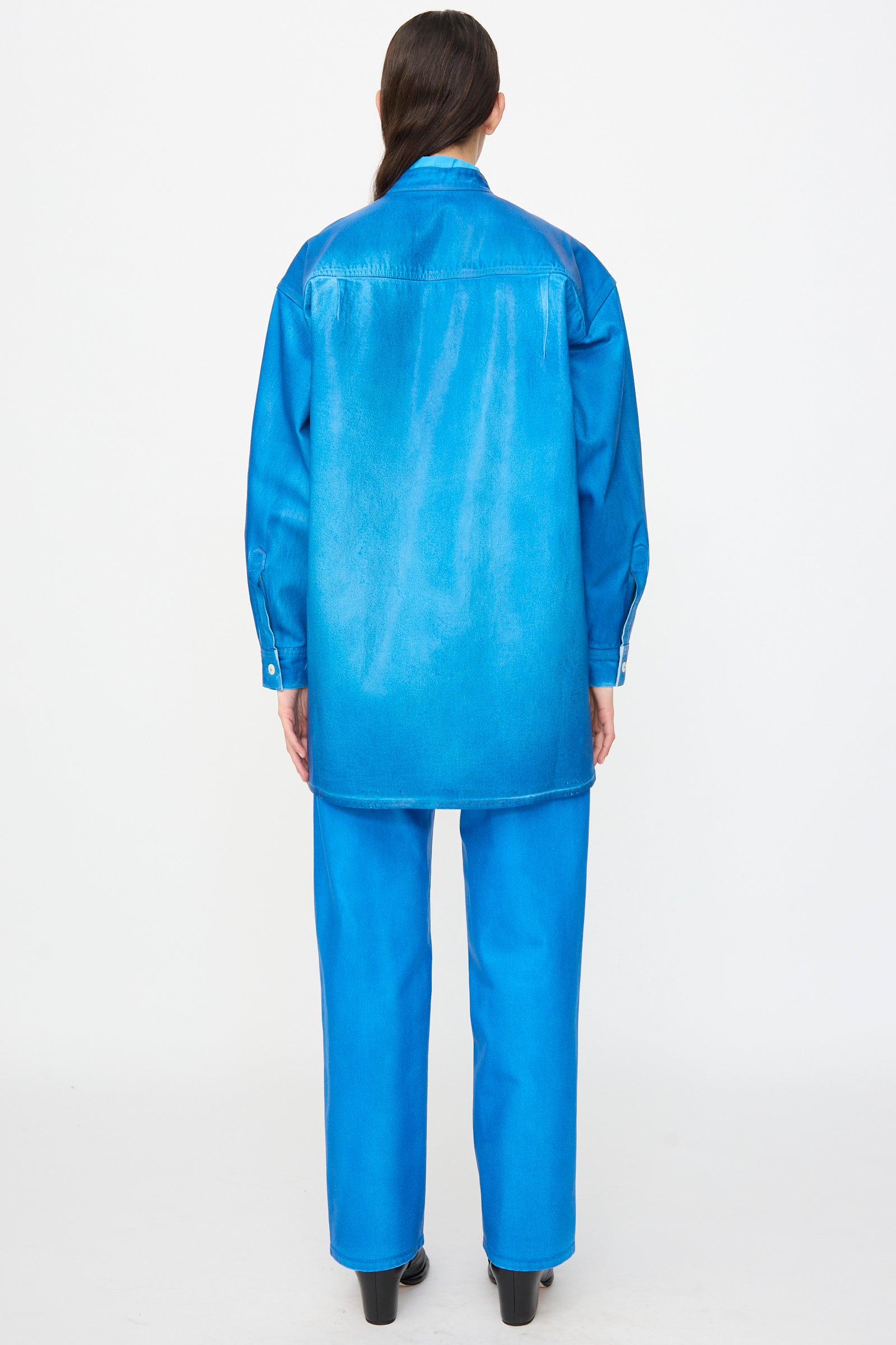 A person with their back to the camera wears the Jesse Kamm Band Collar Okuda Jacket in Electric Blue Lacquer, crafted from regenerative Japanese denim, paired with matching pants, against a plain white background.