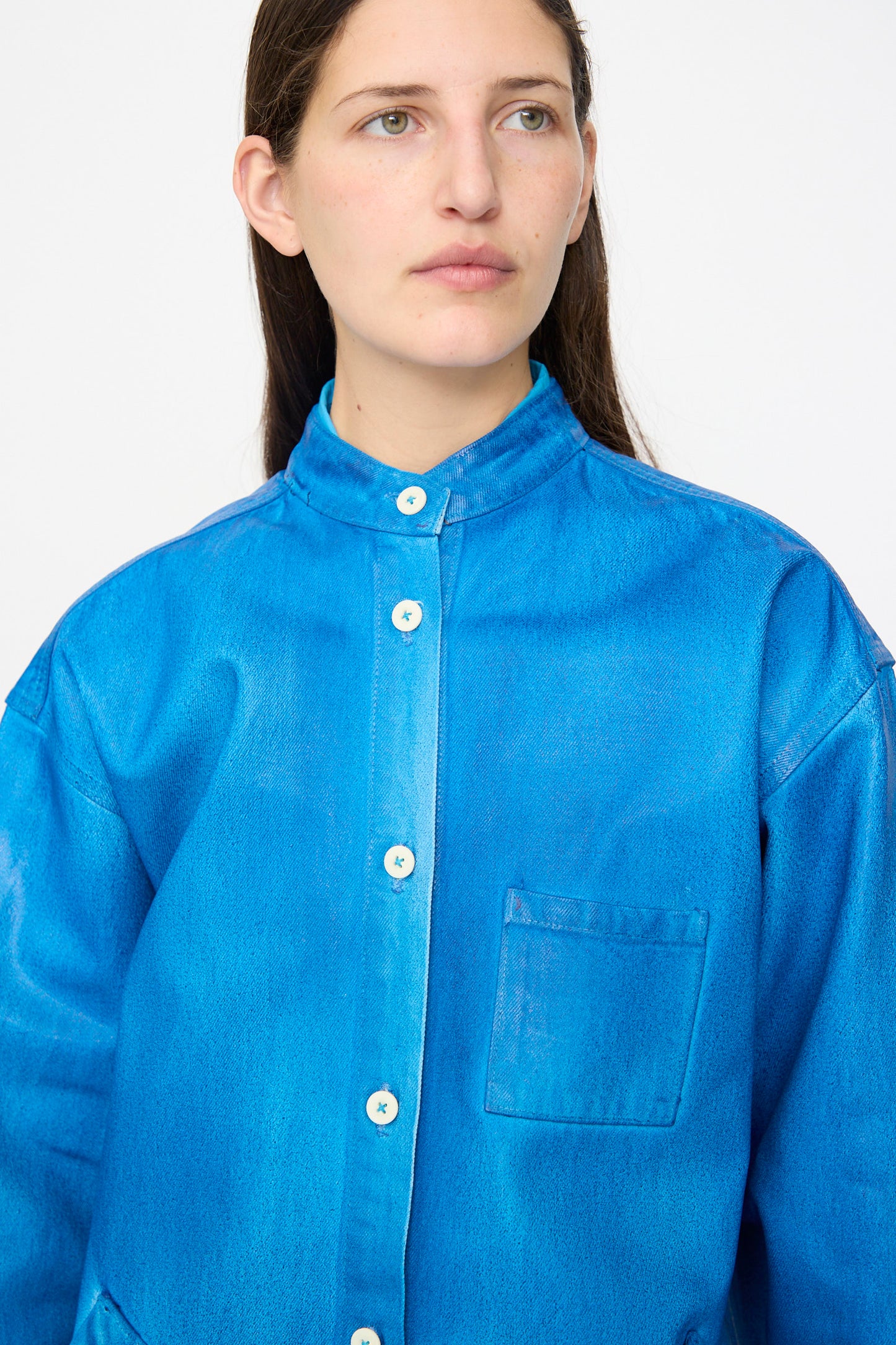 A person with long dark hair wears the Jesse Kamm Band Collar Okuda Jacket in Electric Blue Lacquer, featuring a stand collar and chest pocket, standing against a plain light background.
