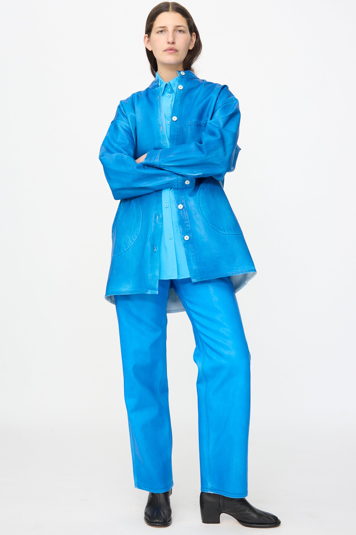 A person stands with arms crossed, wearing the Jesse Kamm Band Collar Okuda Jacket in Electric Blue Lacquer, high-gloss denim pants, and black ankle boots against a plain white background.