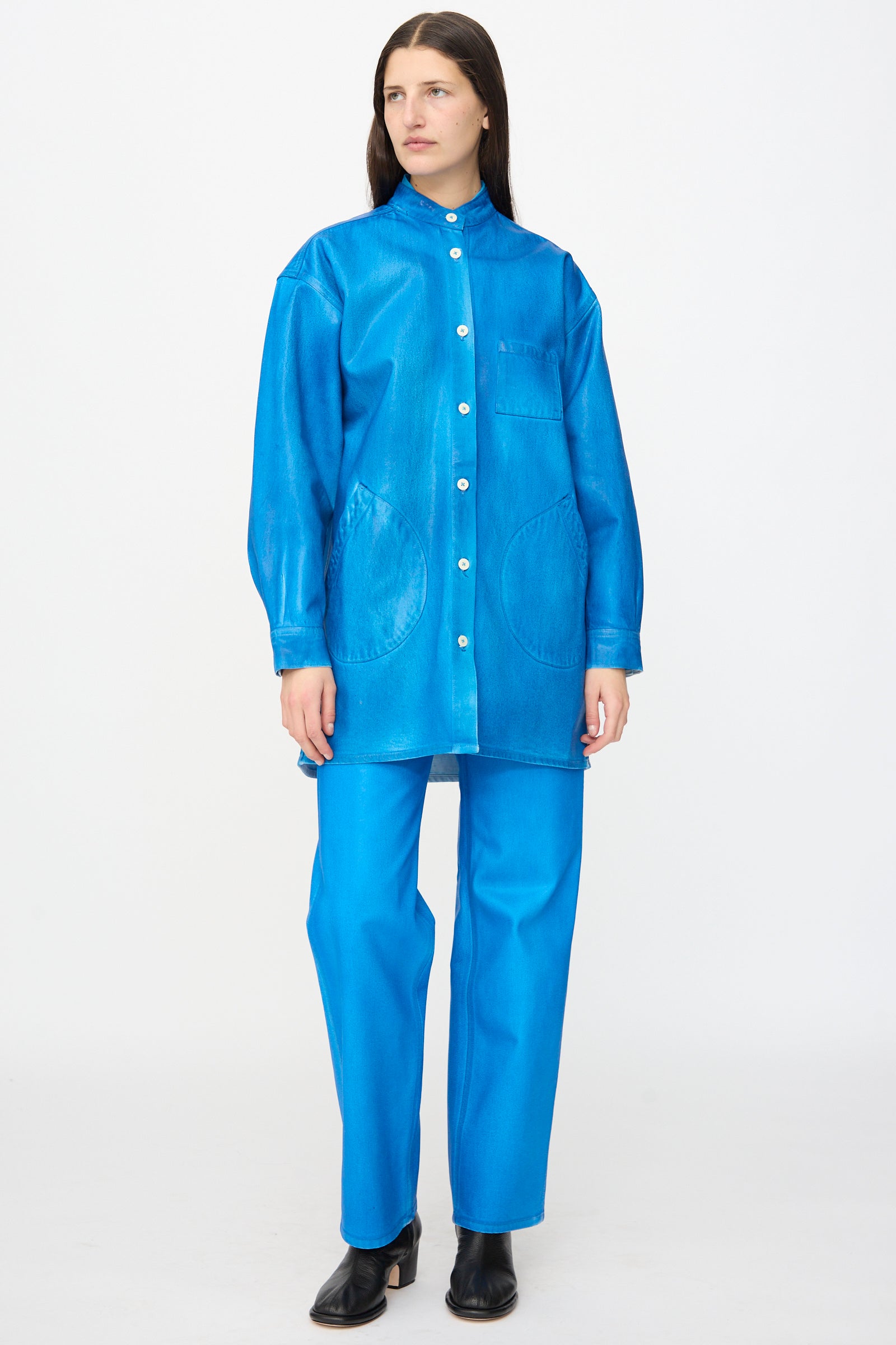 A person wears the Jesse Kamm Band Collar Okuda Jacket in Electric Blue Lacquer, featuring large pockets, paired with matching blue pants and black heeled shoes.