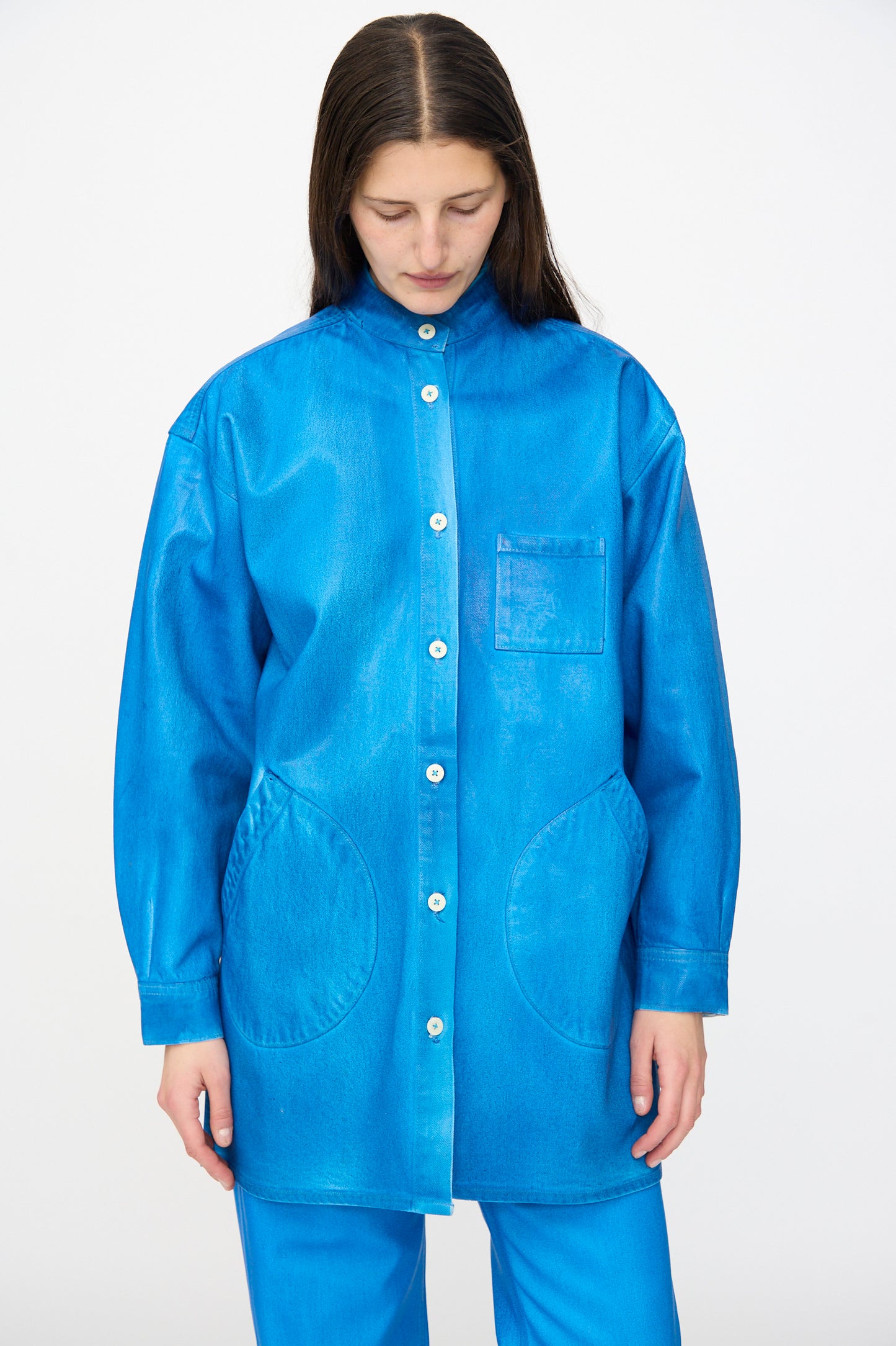 A person with long dark hair looks down, wearing the Jesse Kamm Band Collar Okuda Jacket in Electric Blue Lacquer and matching pants, crafted from regenerative Japanese denim, set against a plain background.
