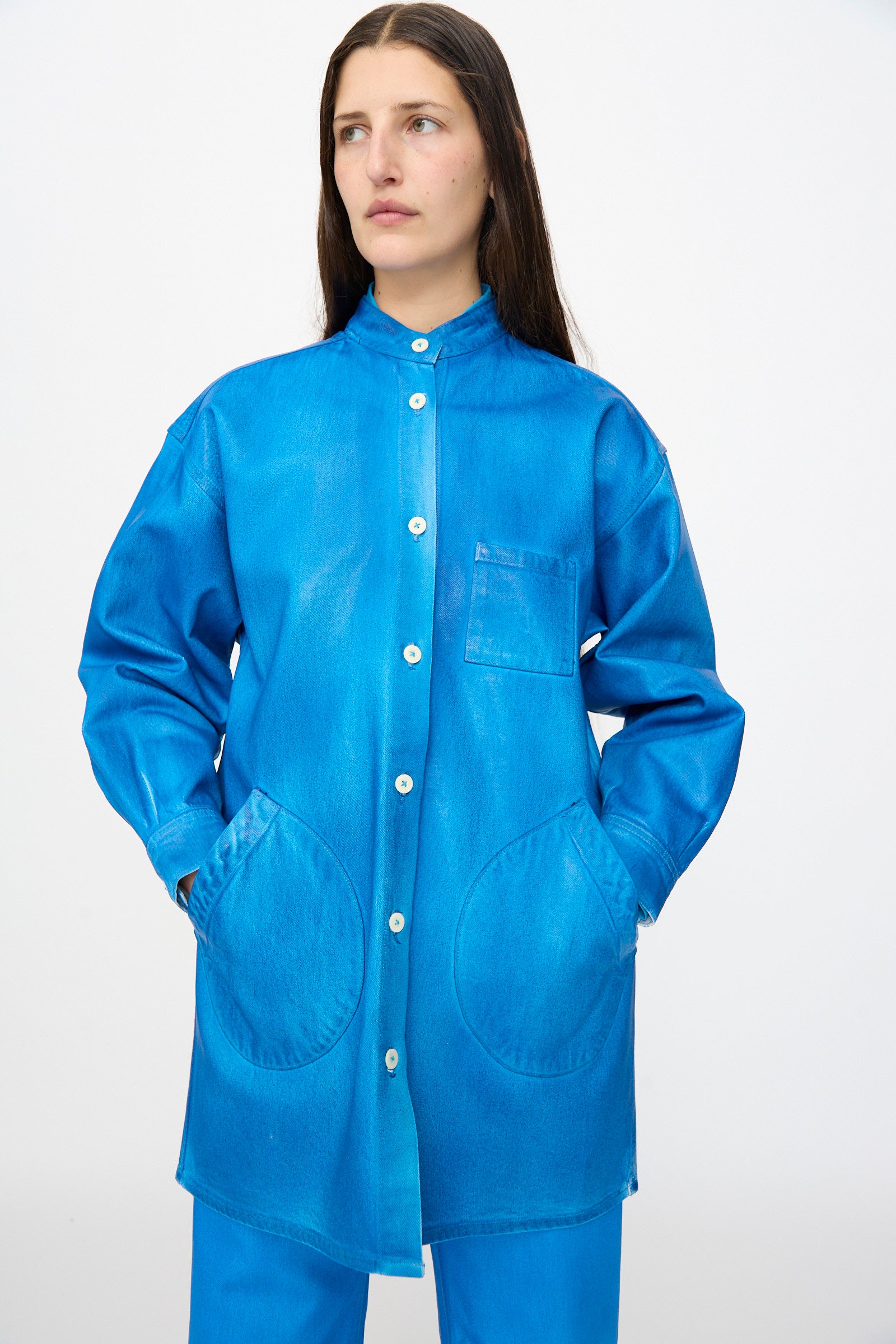 A person with long brown hair wears the Jesse Kamm Band Collar Okuda Jacket in Electric Blue Lacquer, crafted from regenerative Japanese denim, standing with hands in pockets against a plain light background.