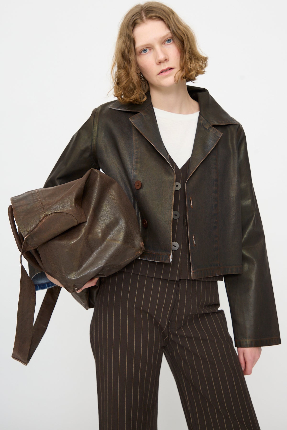 A person stands against a plain white background wearing a dark brown leather jacket, pinstripe pants, and holding the Jesse Kamm Big Bag in Brown Lacquer.