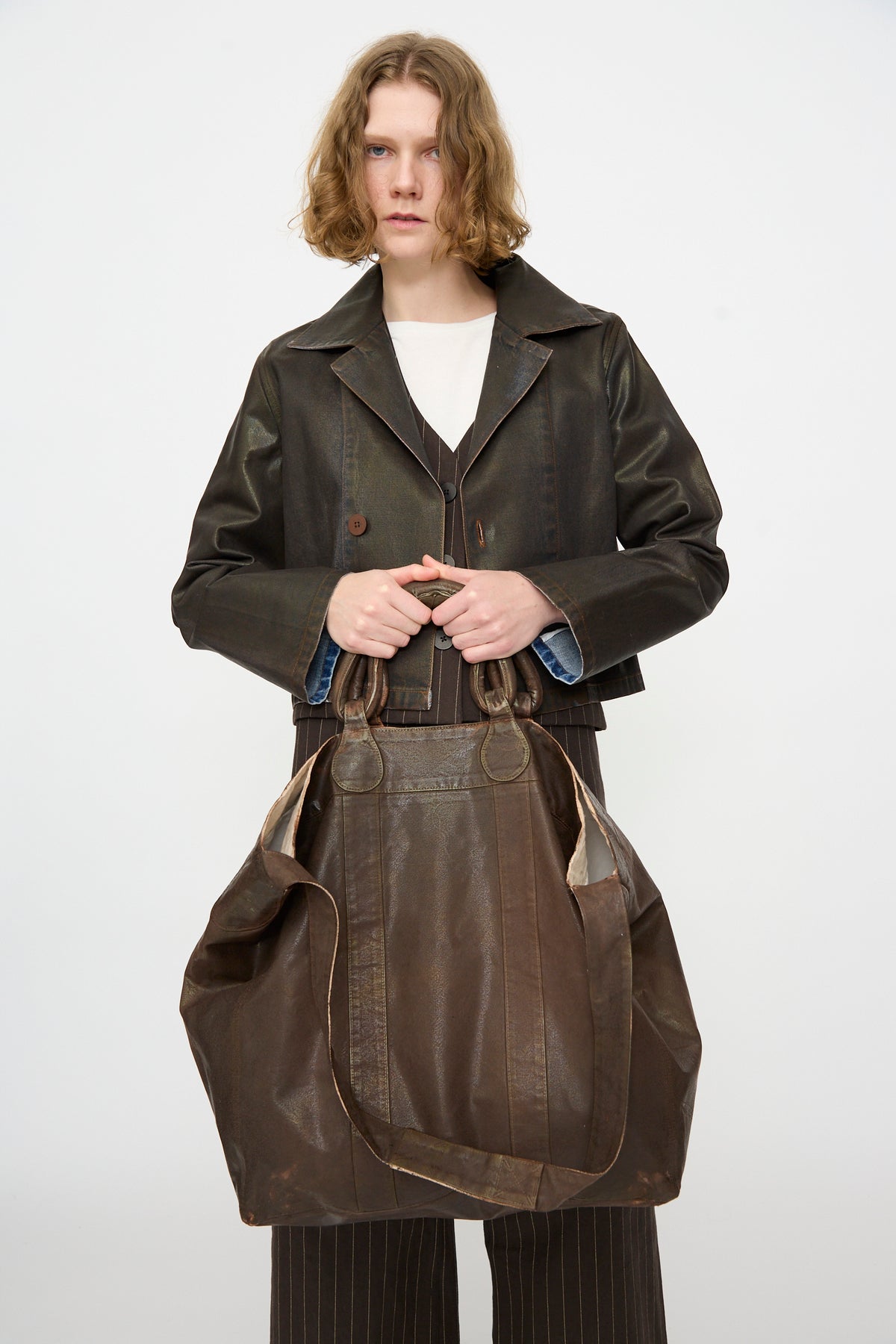 A person with shoulder-length hair, dressed in a dark jacket, holds the Jesse Kamm Big Bag in Brown Lacquer while standing against a plain white background.