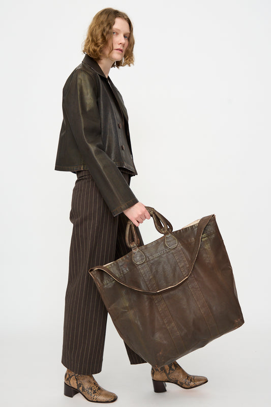 A person holds the Jesse Kamm Big Bag in Brown Lacquer, styled with a dark leather jacket, striped pants, and snakeskin boots, set against a plain white background.