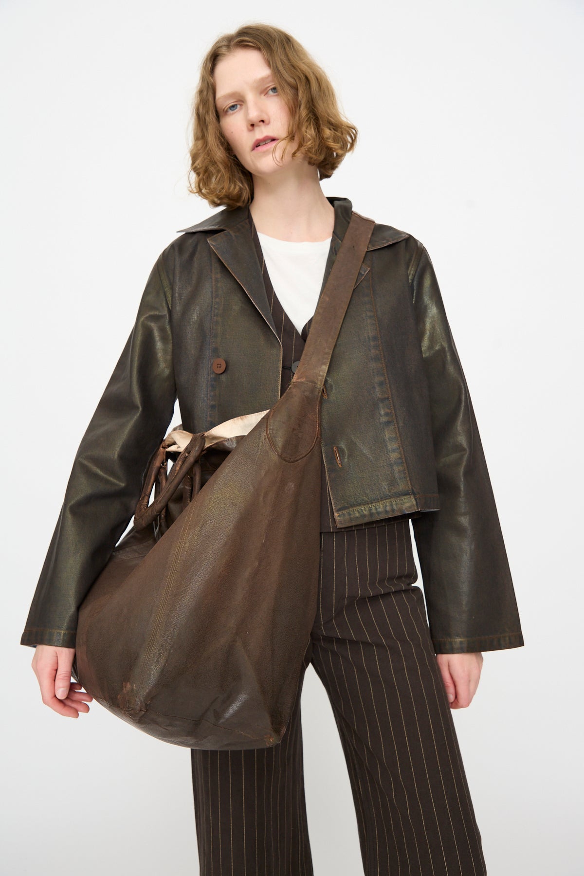 A person with wavy hair wears a dark leather jacket, pinstriped pants, and a crisp white shirt, carrying the Jesse Kamm Big Bag in Brown Lacquer. The background is plain and neutral.