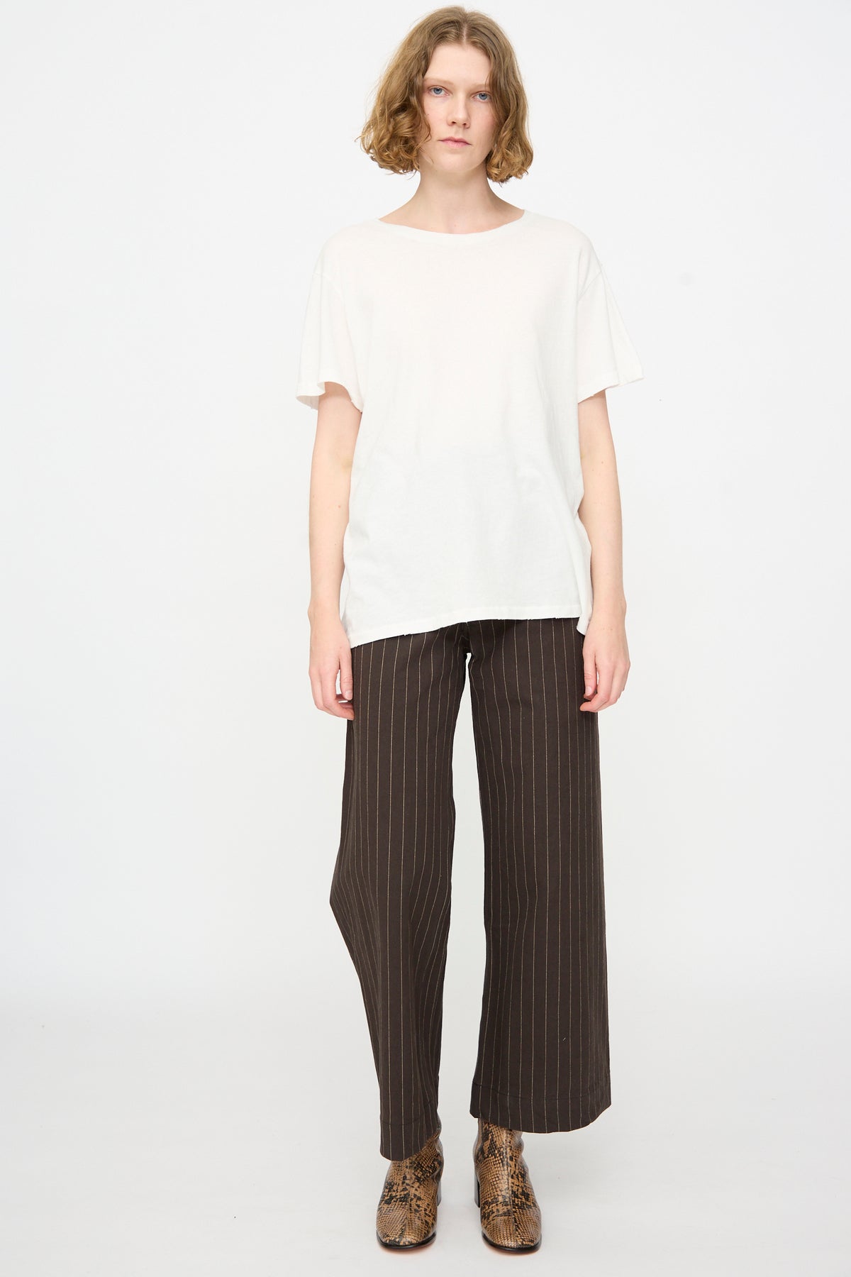 A person stands against a white background wearing the Jesse Kamm Bowery Tee in Salt White, styled with dark wide-leg pinstripe pants and brown snake-print ankle boots.