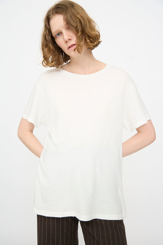 A person with short, wavy, light brown hair wears the Jesse Kamm Bowery Tee in Salt White—a relaxed fit cotton jersey crewneck—paired with dark pinstriped pants, standing against a plain white background.