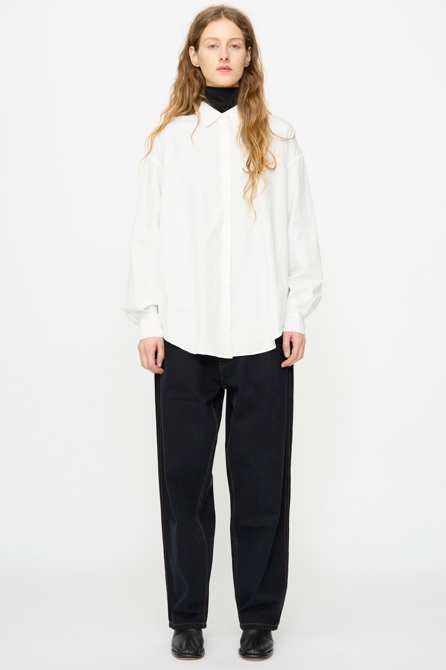 A person stands facing forward against a plain white background, wearing Jesse Kamm's California Wide in Black Overdye denim pants, paired with a white button-up shirt, black turtleneck, and black shoes.