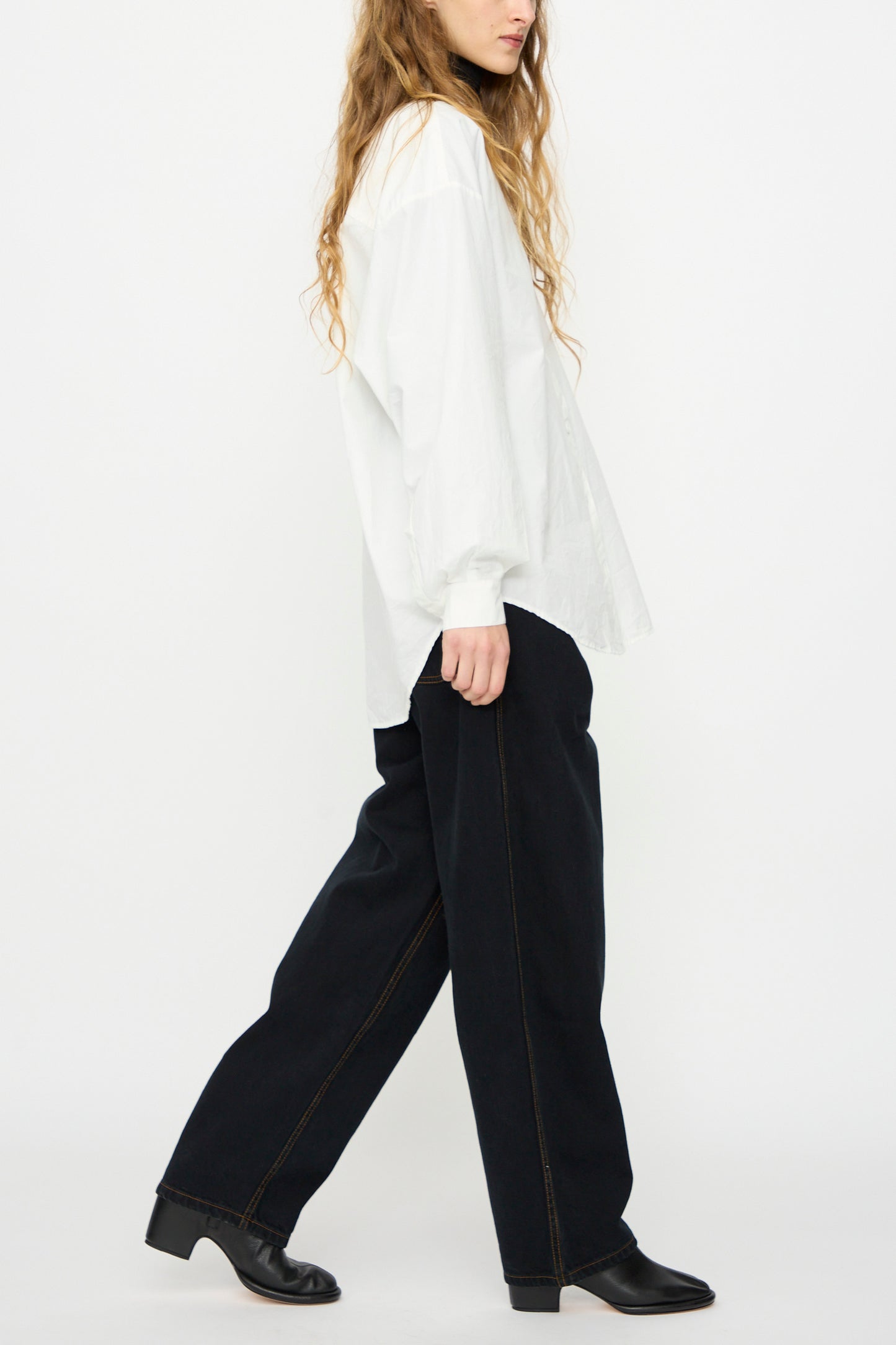 Person in profile wears a loose white shirt, Jesse Kamm's California Wide in Black Overdye unisex organic cotton wide-leg pants, and black shoes against a plain white background.