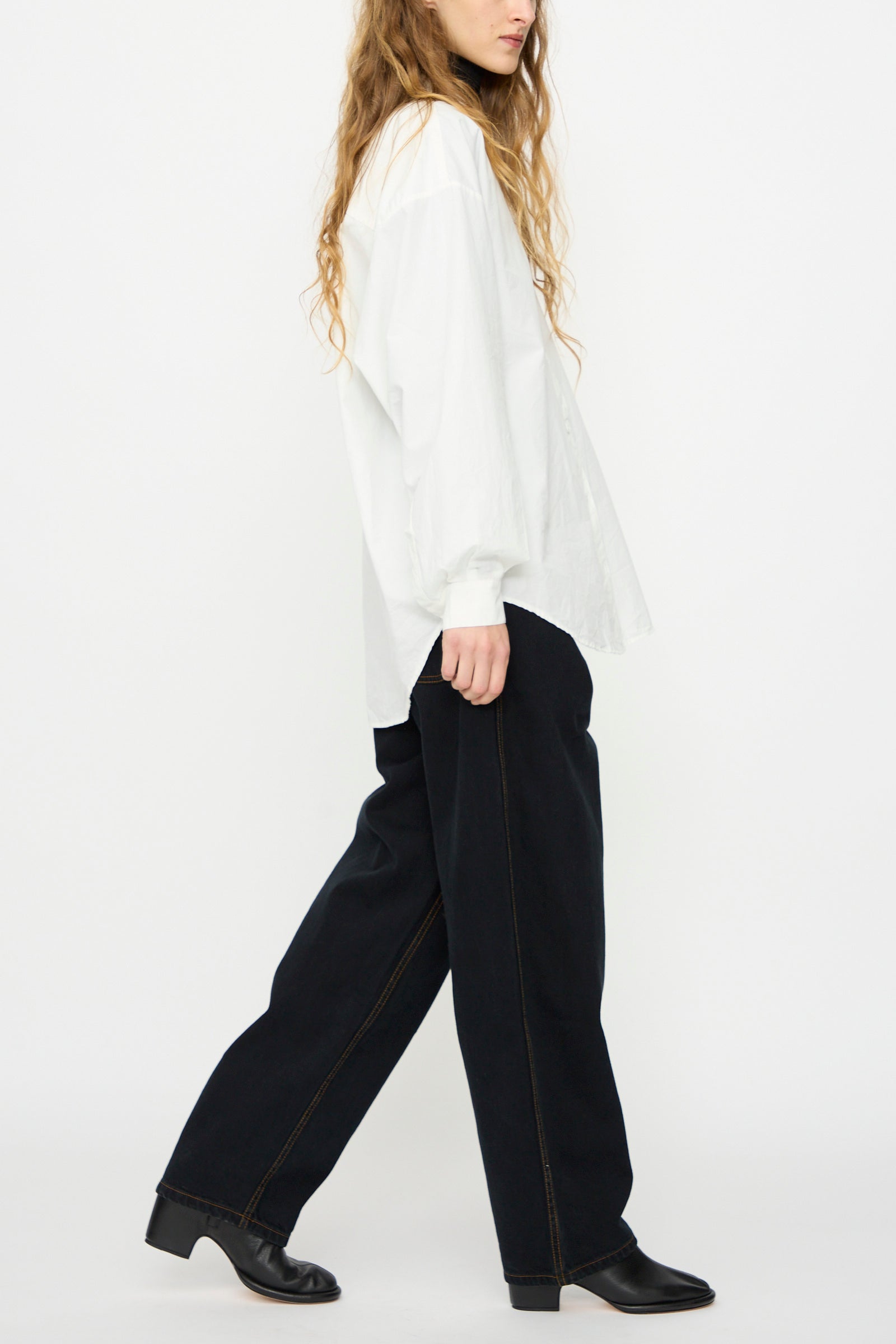 Person in profile wears a loose white shirt, Jesse Kamm's California Wide in Black Overdye unisex organic cotton wide-leg pants, and black shoes against a plain white background.