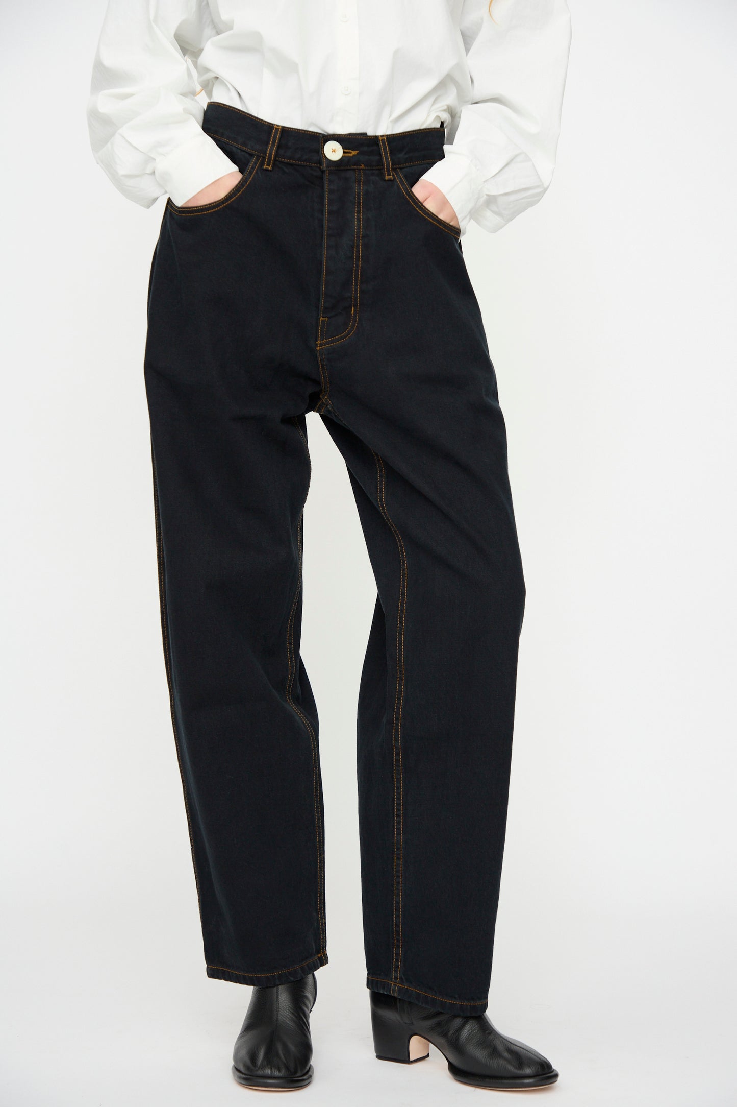 Clad in Jesse Kamm’s California Wide in Black Overdye jeans with orange stitching, a person stands with hands in pockets, wearing a white button-up shirt and black heeled boots.