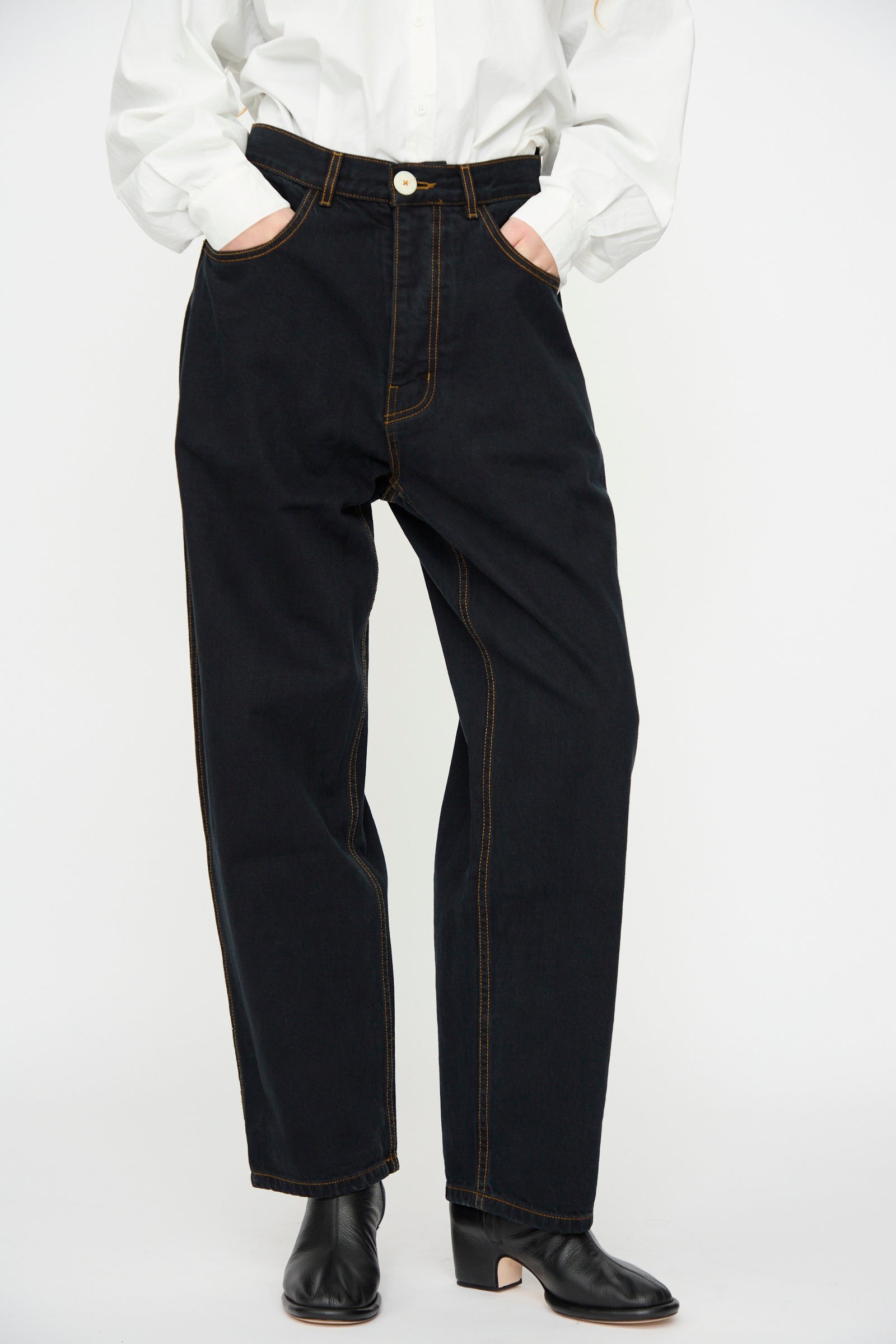 Clad in Jesse Kamm’s California Wide in Black Overdye jeans with orange stitching, a person stands with hands in pockets, wearing a white button-up shirt and black heeled boots.