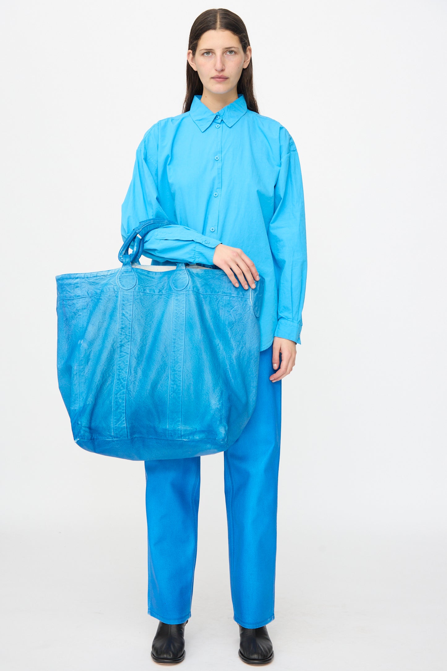 A person stands against a plain background, wearing a bright blue shirt and pants, holding the Jesse Kamm Canvas Big Bag in Electric Blue Lacquer.