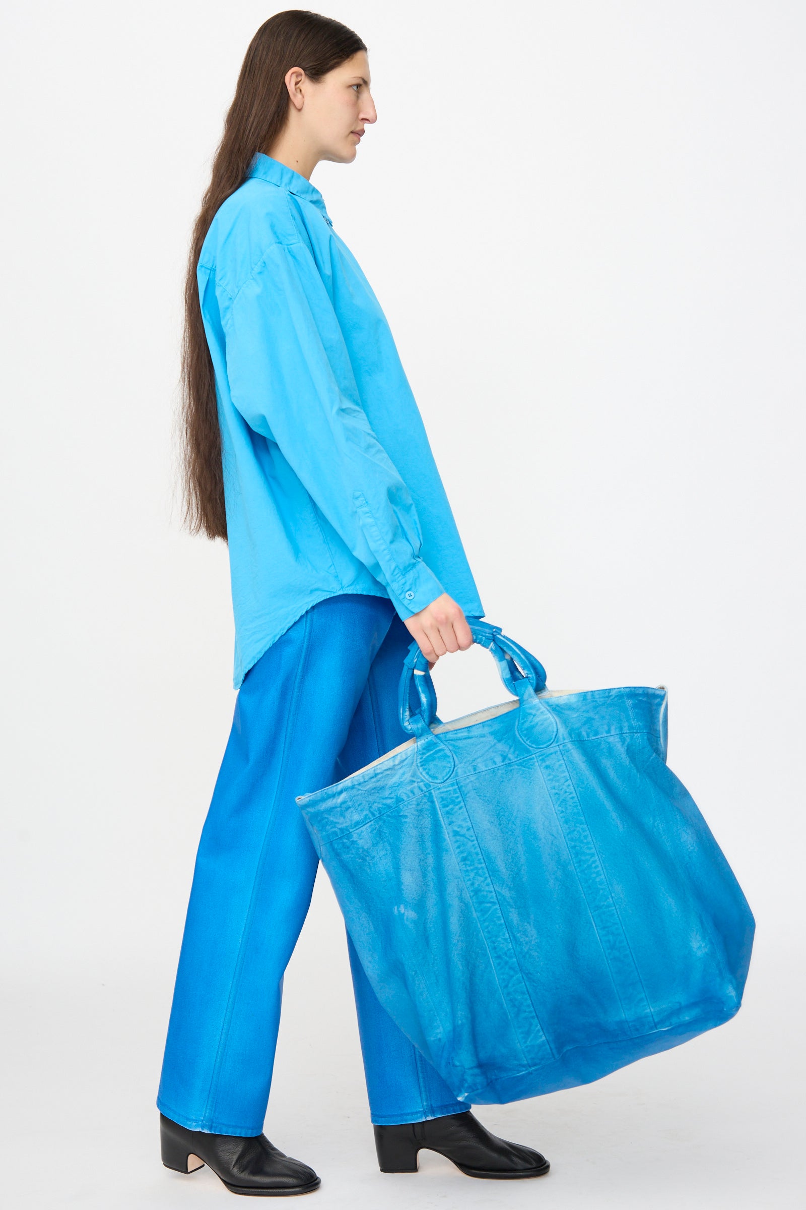 A person in a loose blue shirt and pants holds the Jesse Kamm Canvas Big Bag in Electric Blue Lacquer, standing sideways against a plain white background.