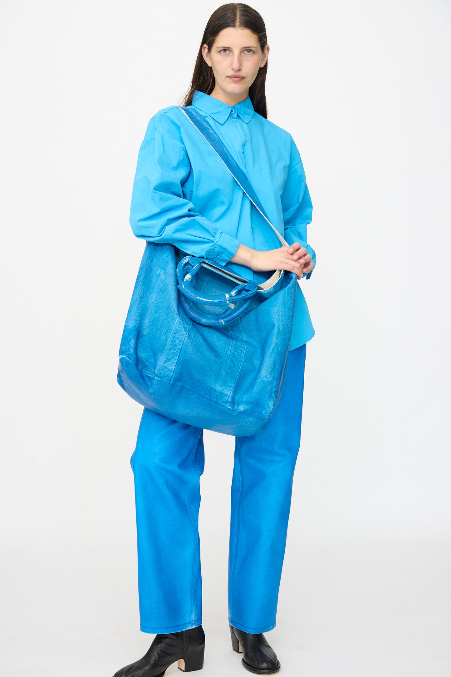 A person stands against a plain background wearing a bright blue shirt and pants, holding the Jesse Kamm Canvas Big Bag in Electric Blue Lacquer, and wearing black ankle boots.