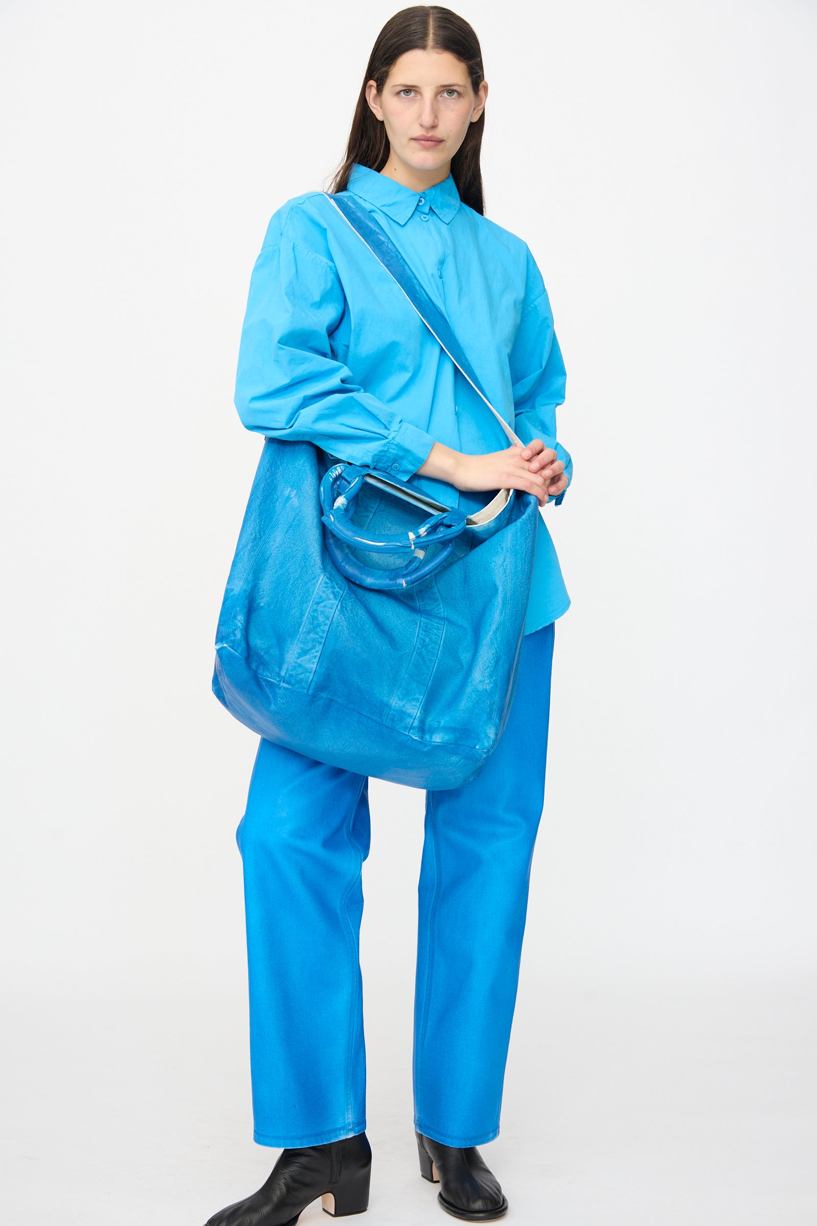 A person stands against a plain background wearing a bright blue shirt and pants, holding the Jesse Kamm Canvas Big Bag in Electric Blue Lacquer, and wearing black ankle boots.